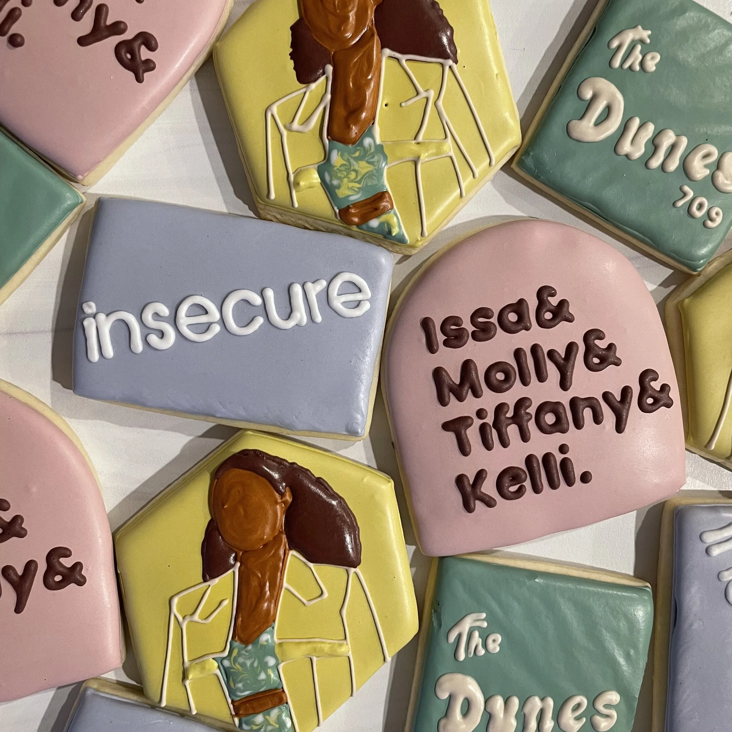 Decorative cookies with colorful pastel icing, some have words like 'Insecurity' and 'The D**e' written in white icing, and some feature abstract designs. The cookies are arranged in a close group.