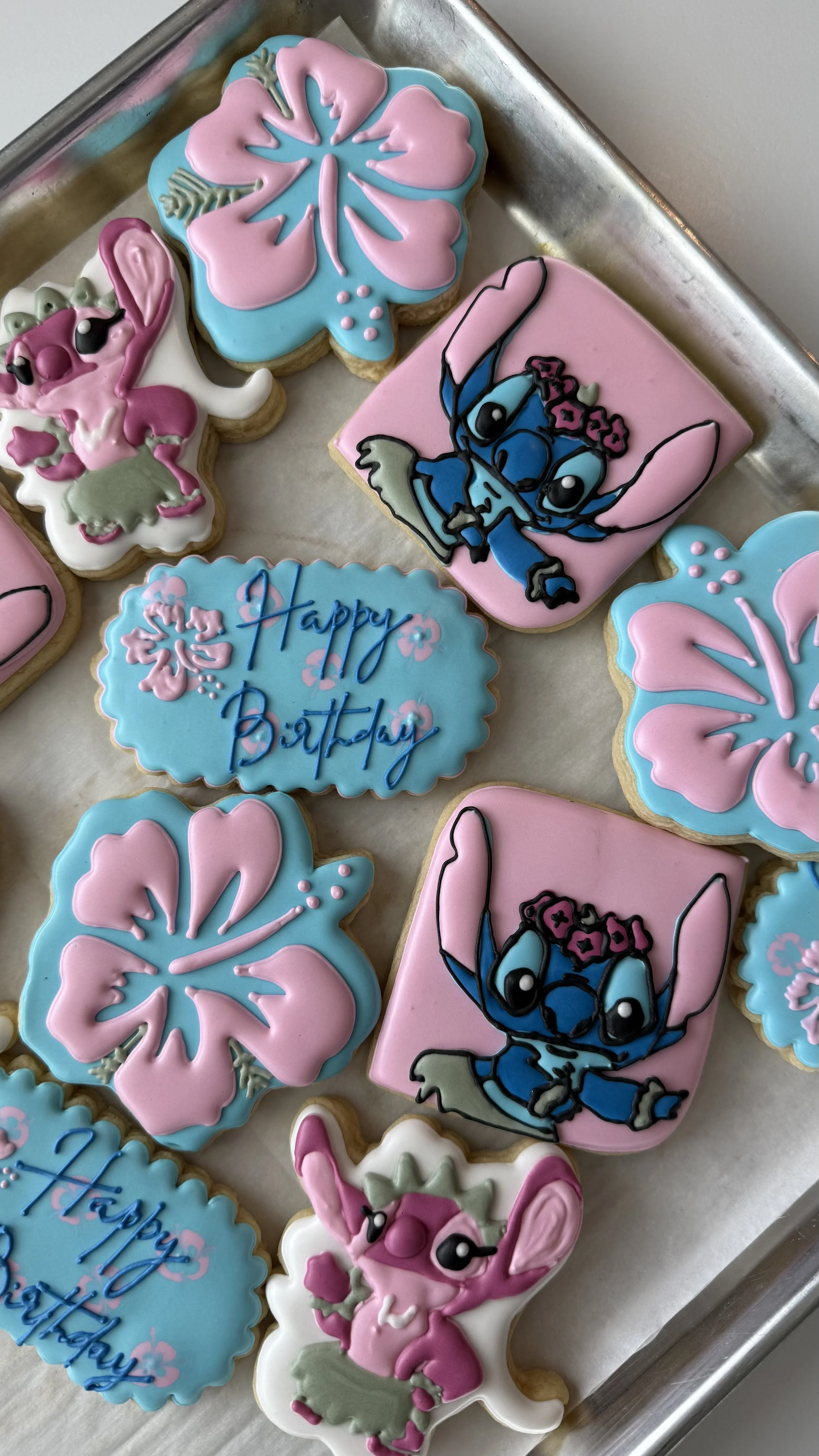 Decorated cookies with pink and blue icing, featuring images of a hibiscus flower, the character Stitch from Lilo & Stitch, and a bunny, along with a cookie that says "Happy Birthday."