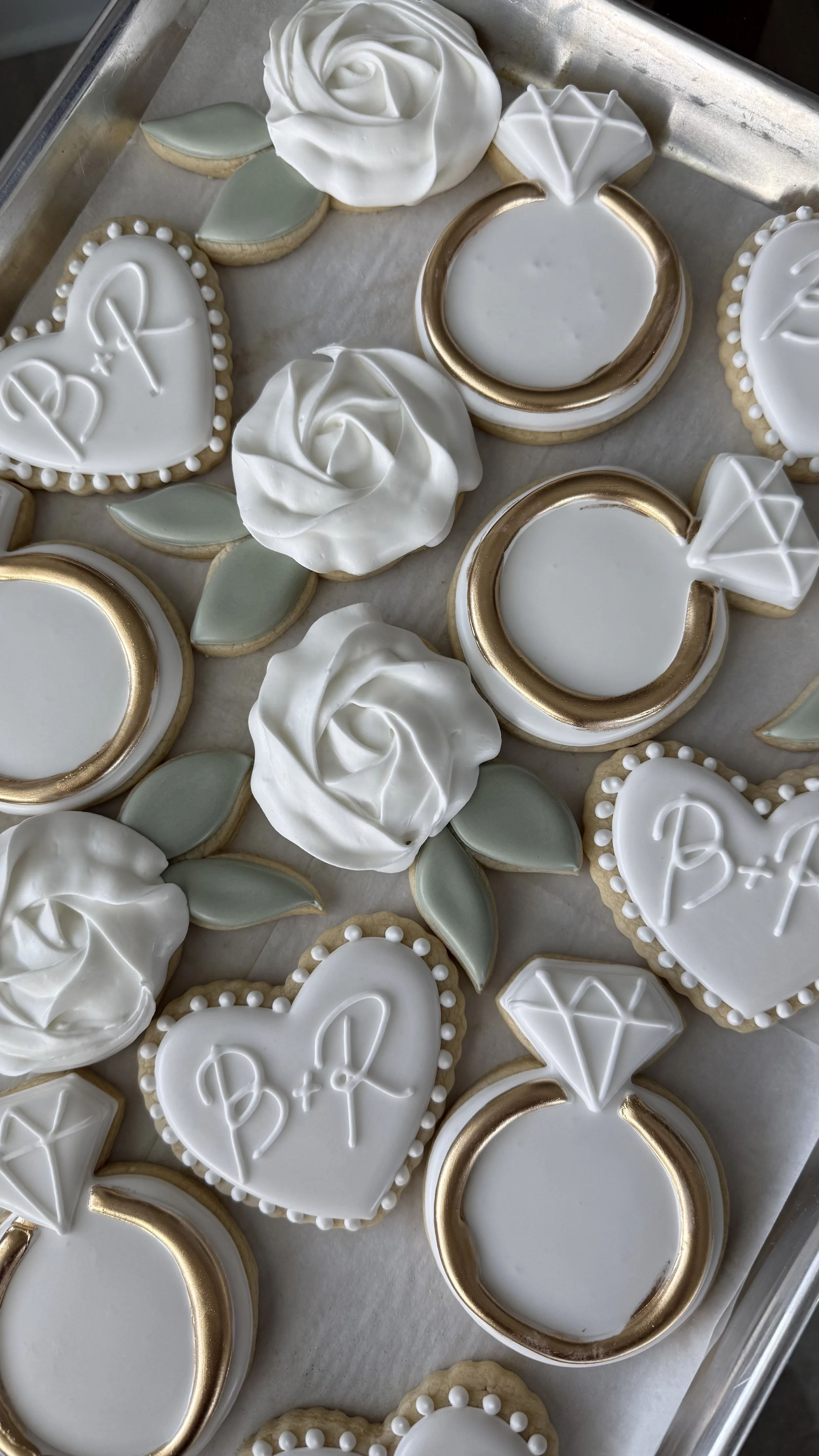 Elegant cookies decorated for a wedding or engagement, featuring white icing, gold accents, rose designs, leaf shapes, and diamond motifs.