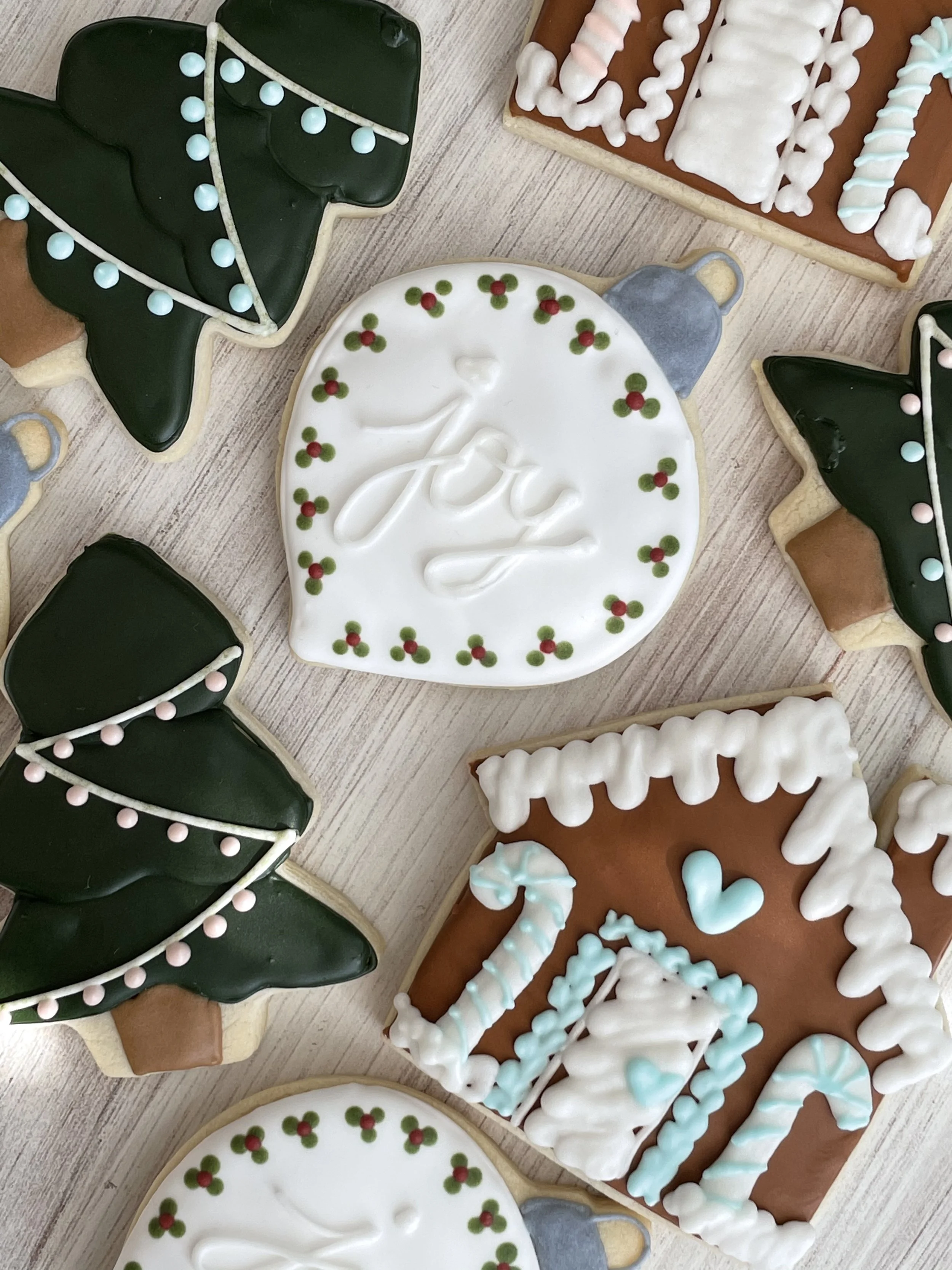 Several decorated Christmas cookies including Christmas trees with dark green icing and pink and blue decorations, a white round cookie with holiday holly decoration and white piping, and rectangular cookies with white and blue icing stripes.