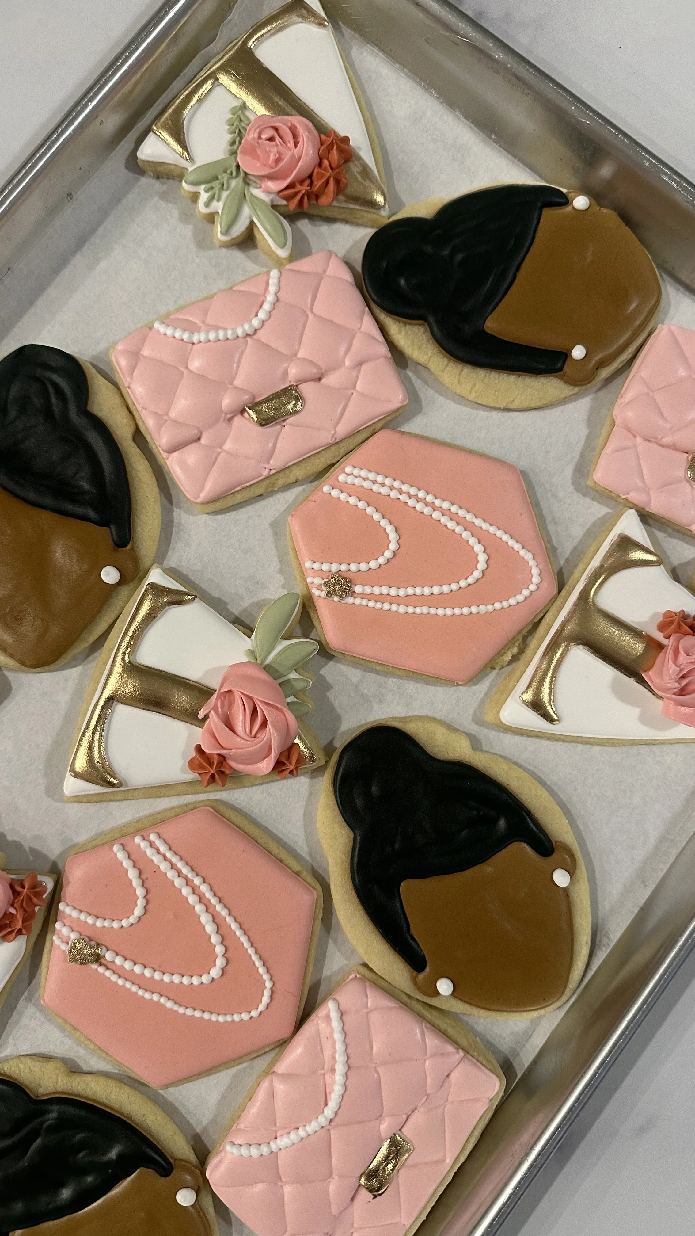 Decorative cookies with various designs, including pink and white quilted patterns with pearl-like accents, black and brown hair-themed cookies, and letter 'F' cookies with floral and pearl decorations, on a silver tray.