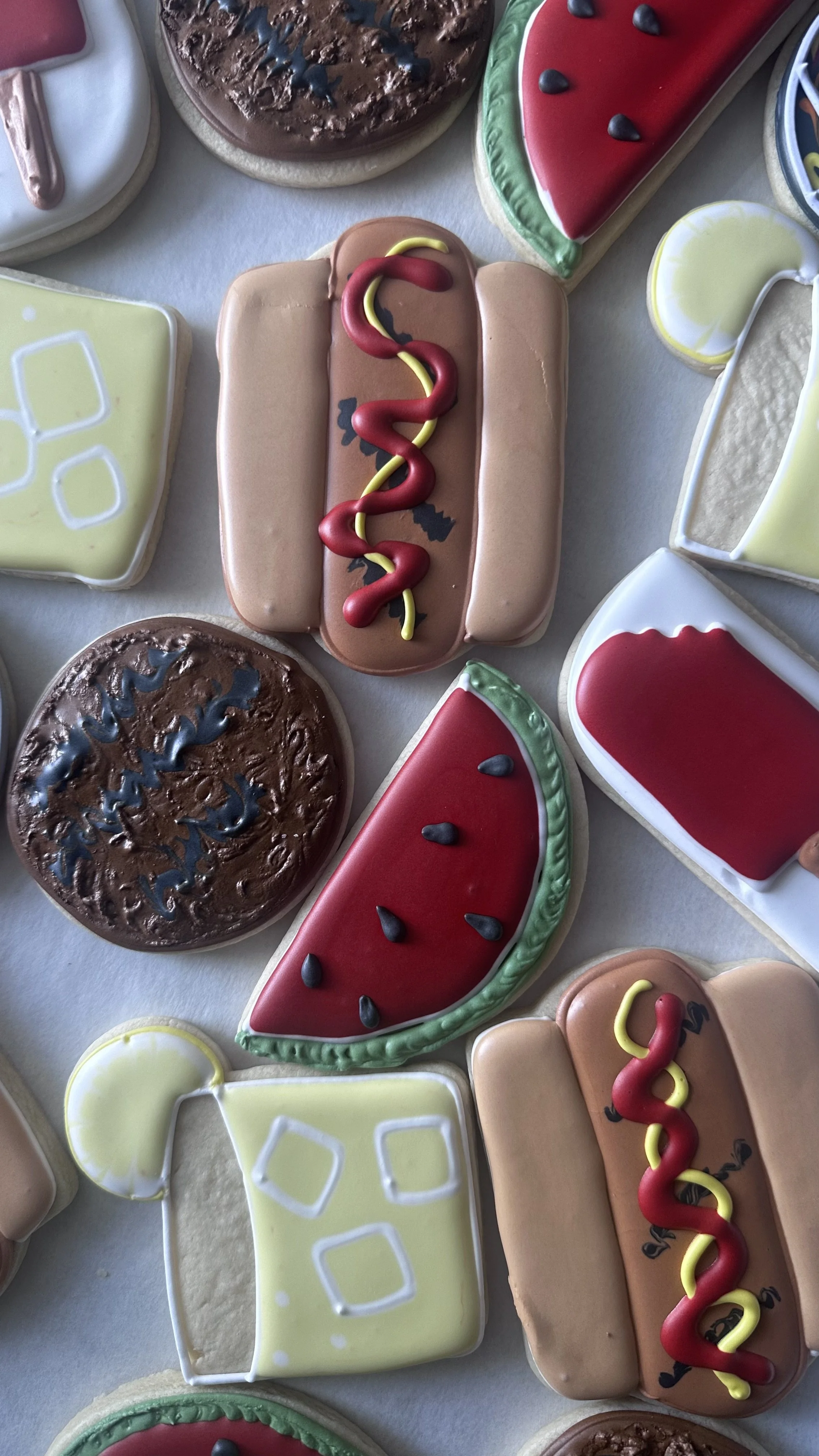 Assorted decorated cookies shaped and decorated like various food items, including a hot dog, pizza slices, watermelon slices, a lemonade glass, and other food-themed designs.