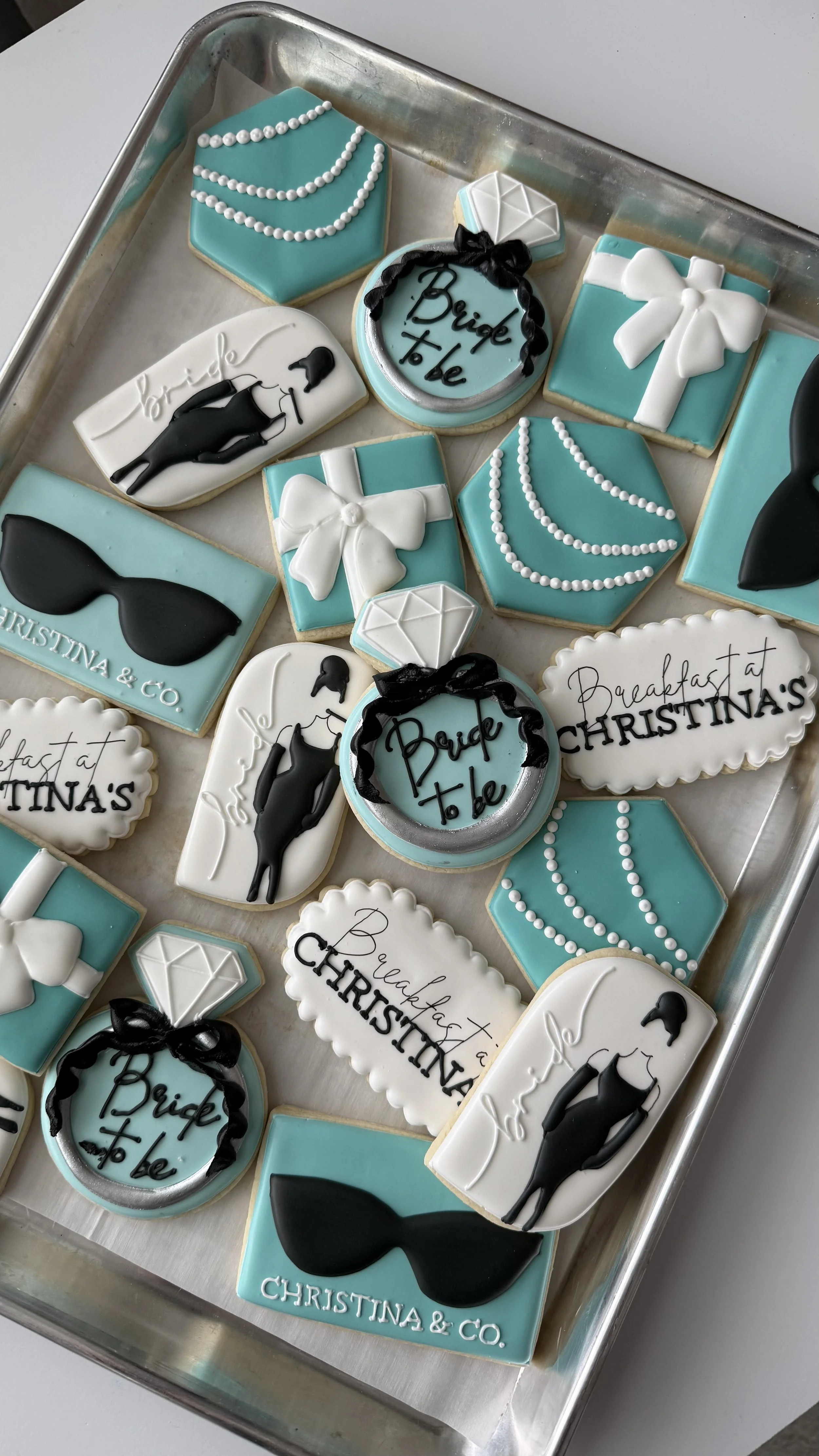 Assorted cookies with bridal and wedding themes, decorated with white, black, and blue icing, featuring phrases like 'Bride to be' and 'Breakfast at Christina's,' along with images of sunglasses, diamonds, bows, and a woman in a dress.