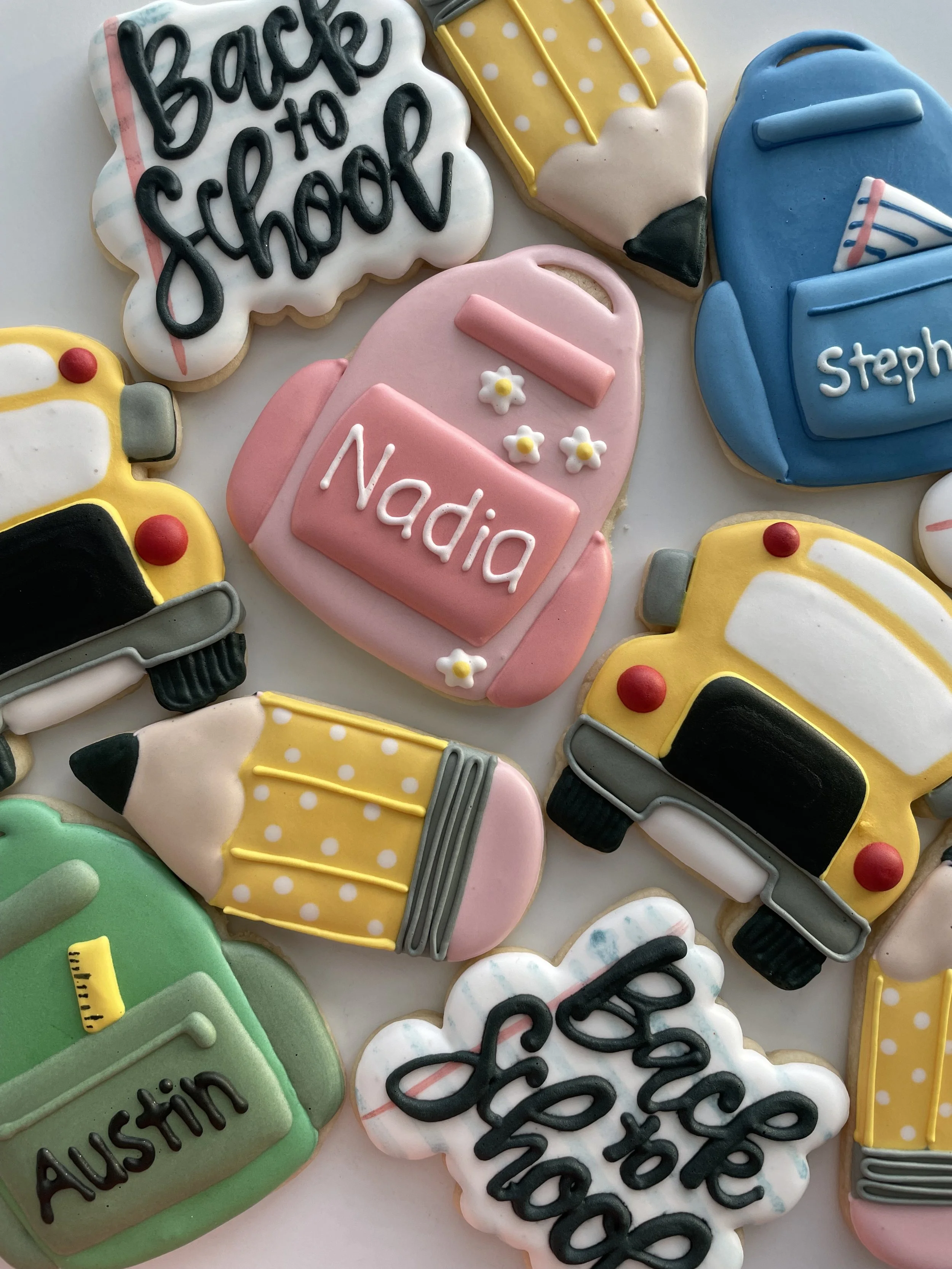 Decorated sugar cookies in various shapes including a backpack with the name 'Steph', a yellow school bus, a pencil, and a cloud-shaped cookie with the words 'Back to School' written on it. The cookies are colorful with icing and embellishments.