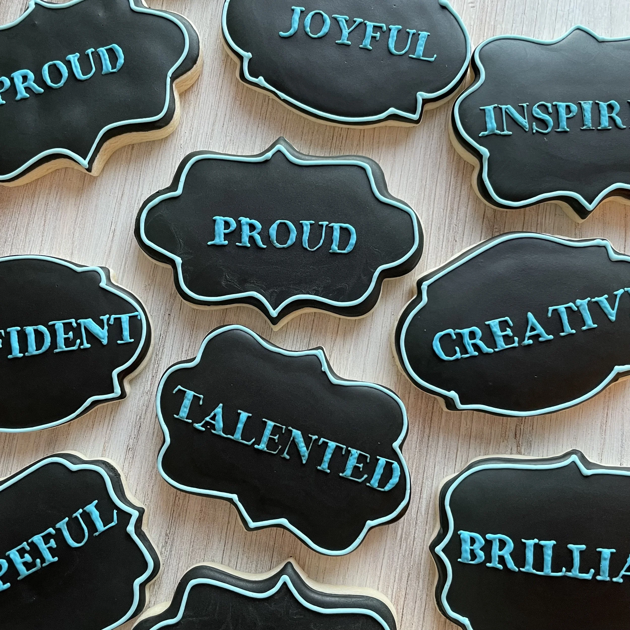 Decorative cookies with words like 'Proud,' 'Joyful,' and 'Inspire' written in blue icing on a black background with a light border, shaped like speech bubbles.
