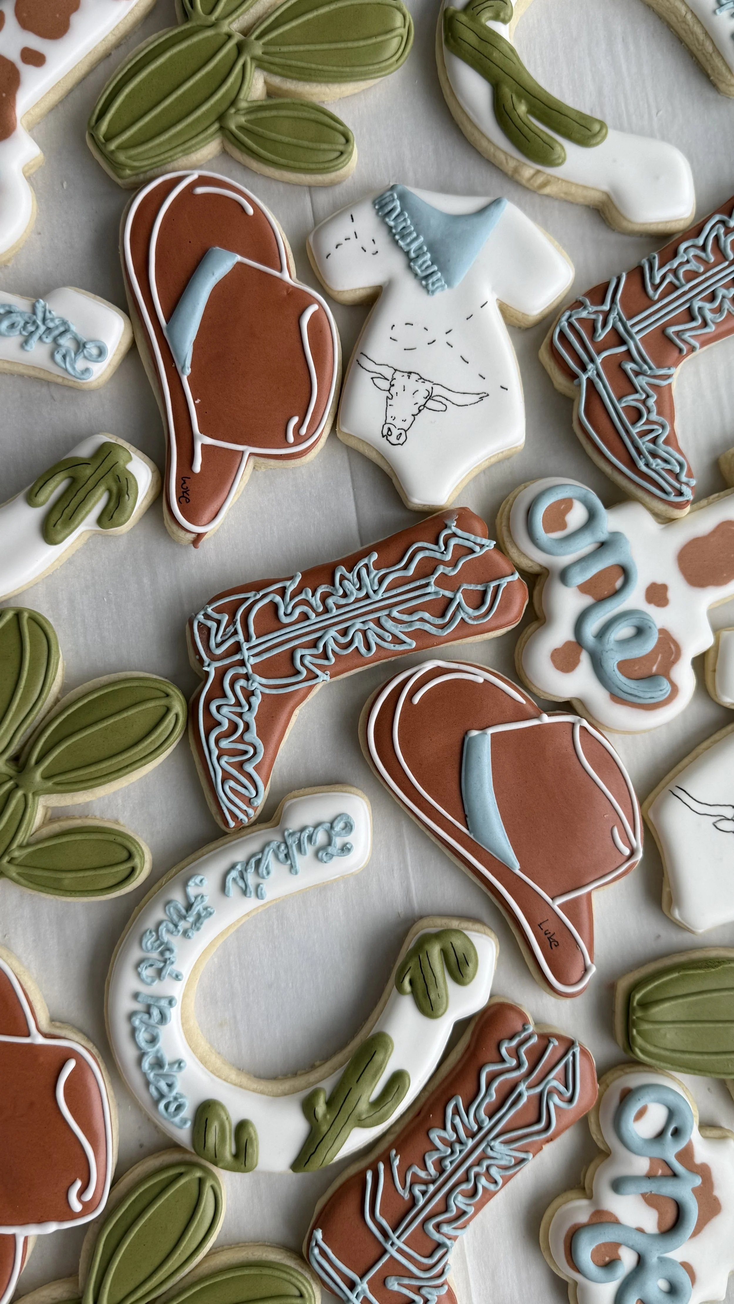 Decorated cookies in desert-themed shapes, including cactus, cowboy hat, roping saddle, and horse wearing a blue hat.