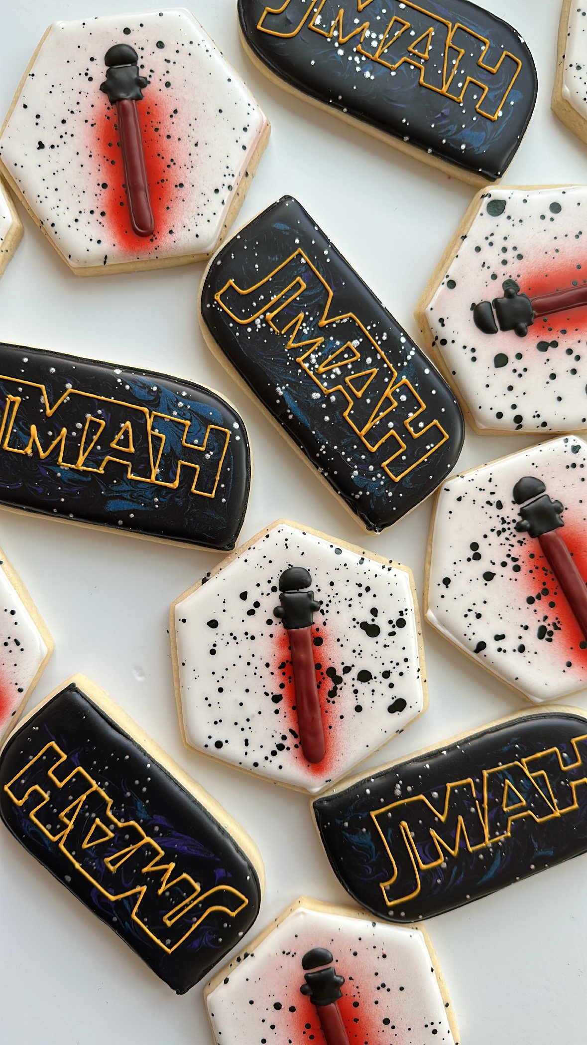 Halloween-themed cookies decorated with black, white, and dark blue icing, some featuring word art and others with a black figure resembling a ghost or skeleton attached to a red stick, arranged on a white surface.