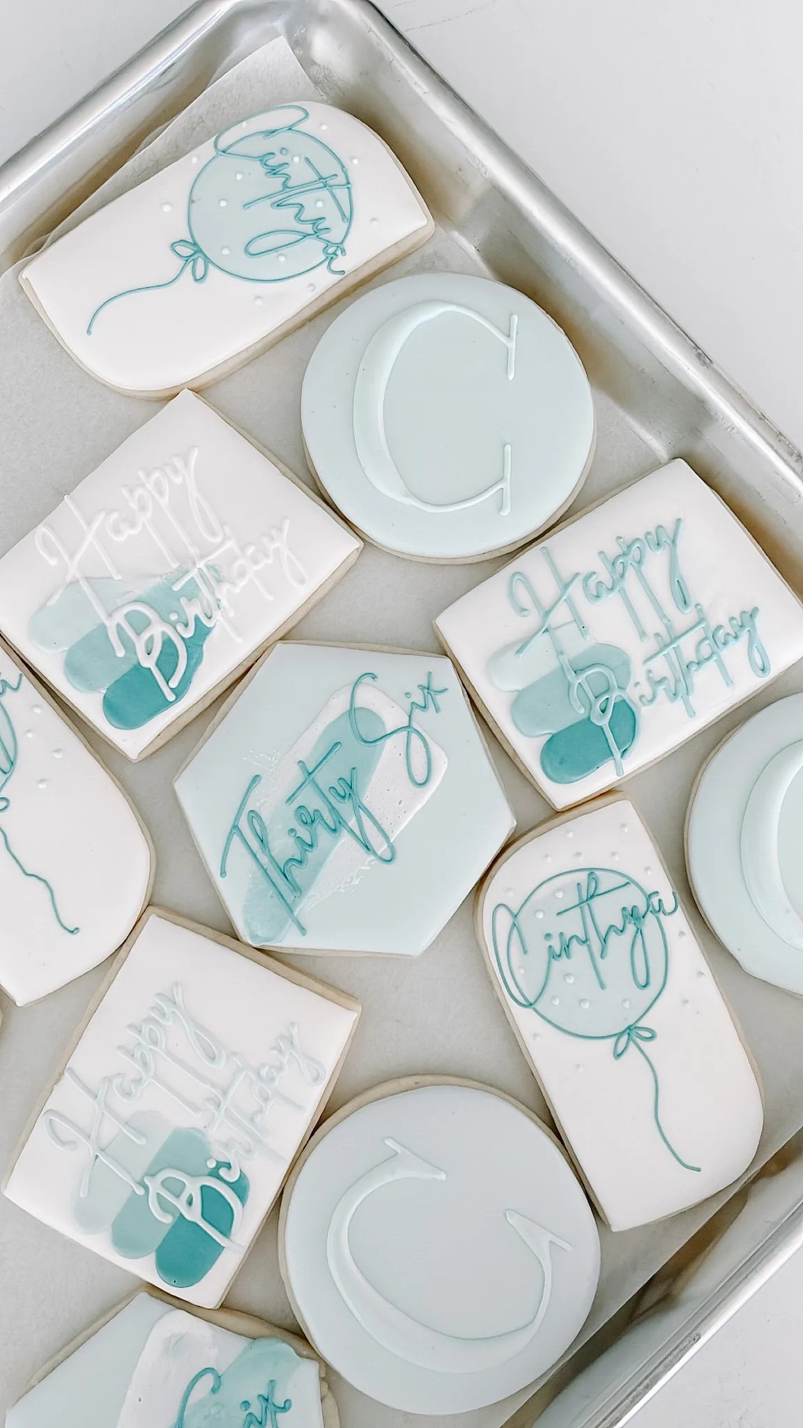 Assorted white cookies with light blue icing, some with 'happy birthday' written and decorative balloon drawings.