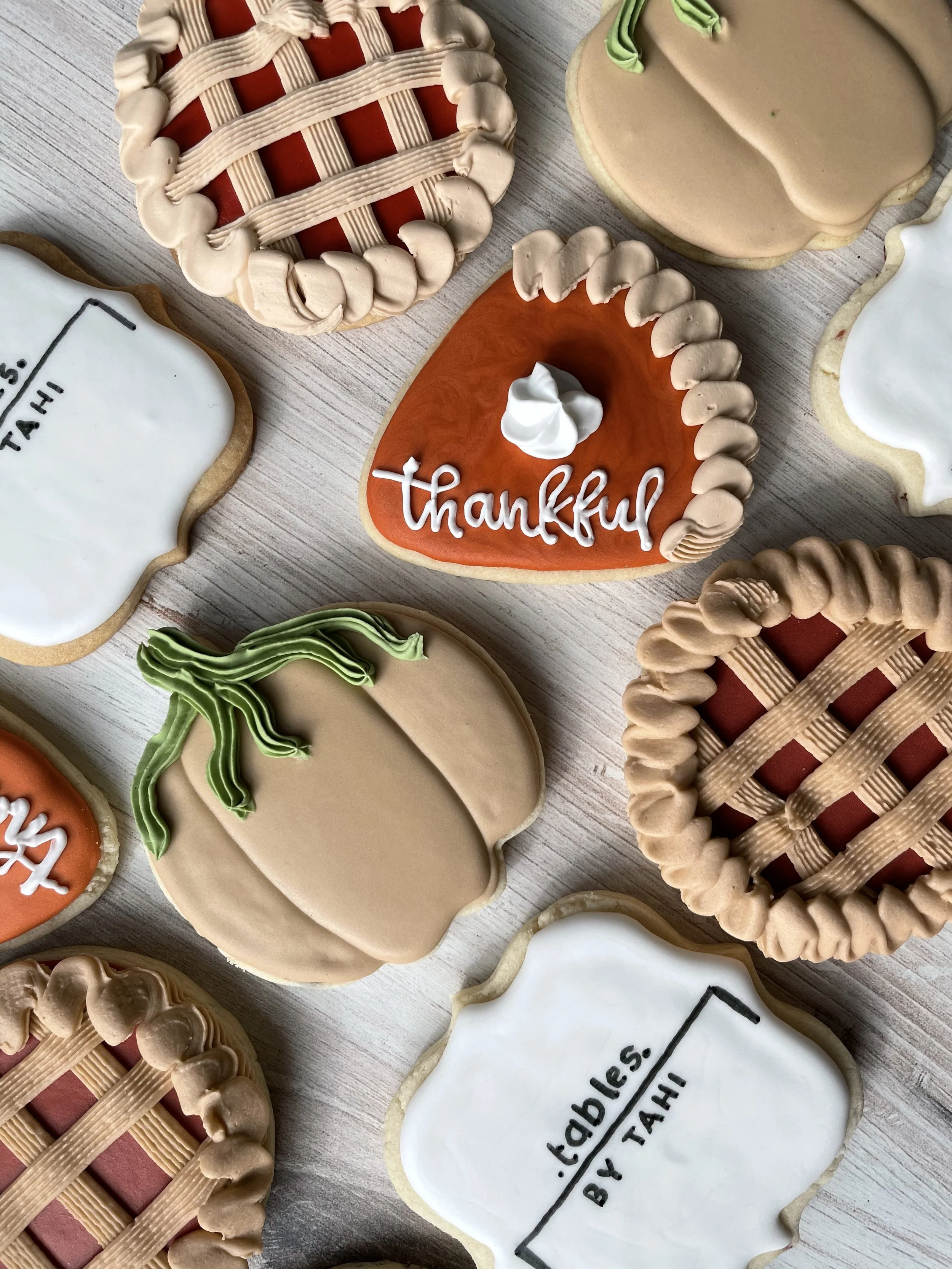 A variety of decorated cookies, including 'thankful' and graduation-themed cookies, decorated with frosting and intricate icing designs, laid out on a light wooden surface.