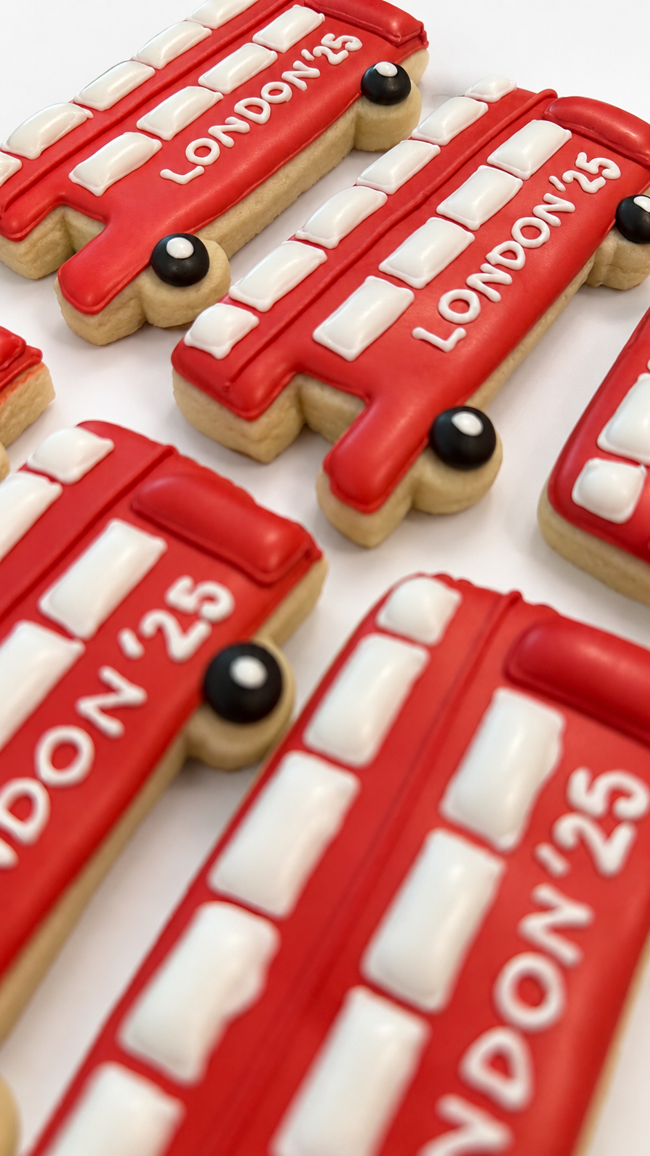 Decorative cookies shaped and decorated like London buses with red icing, white windows, black wheels, and the text "LONDON '25" written on them.