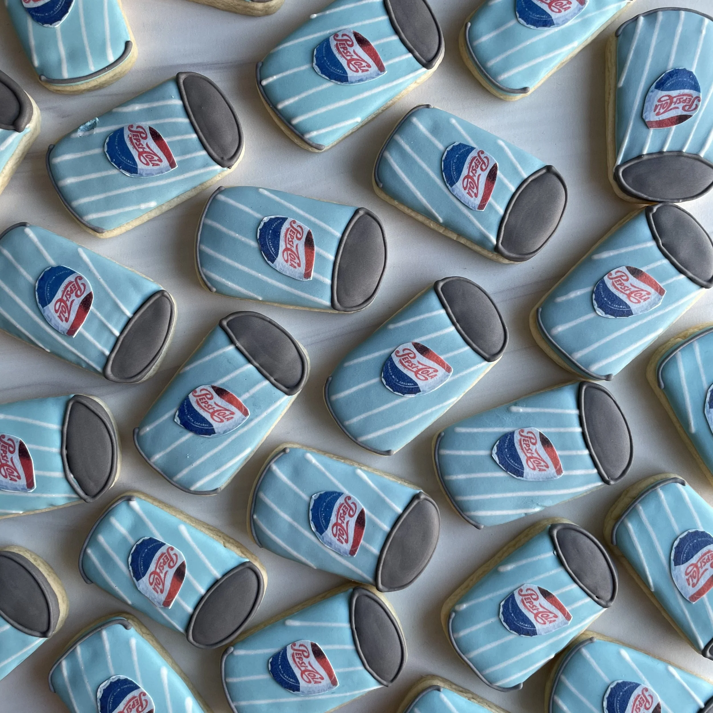 A close-up of rectangular cookies decorated with blue icing, white stripes, a Pepsi logo, and black details resembling sunglasses.