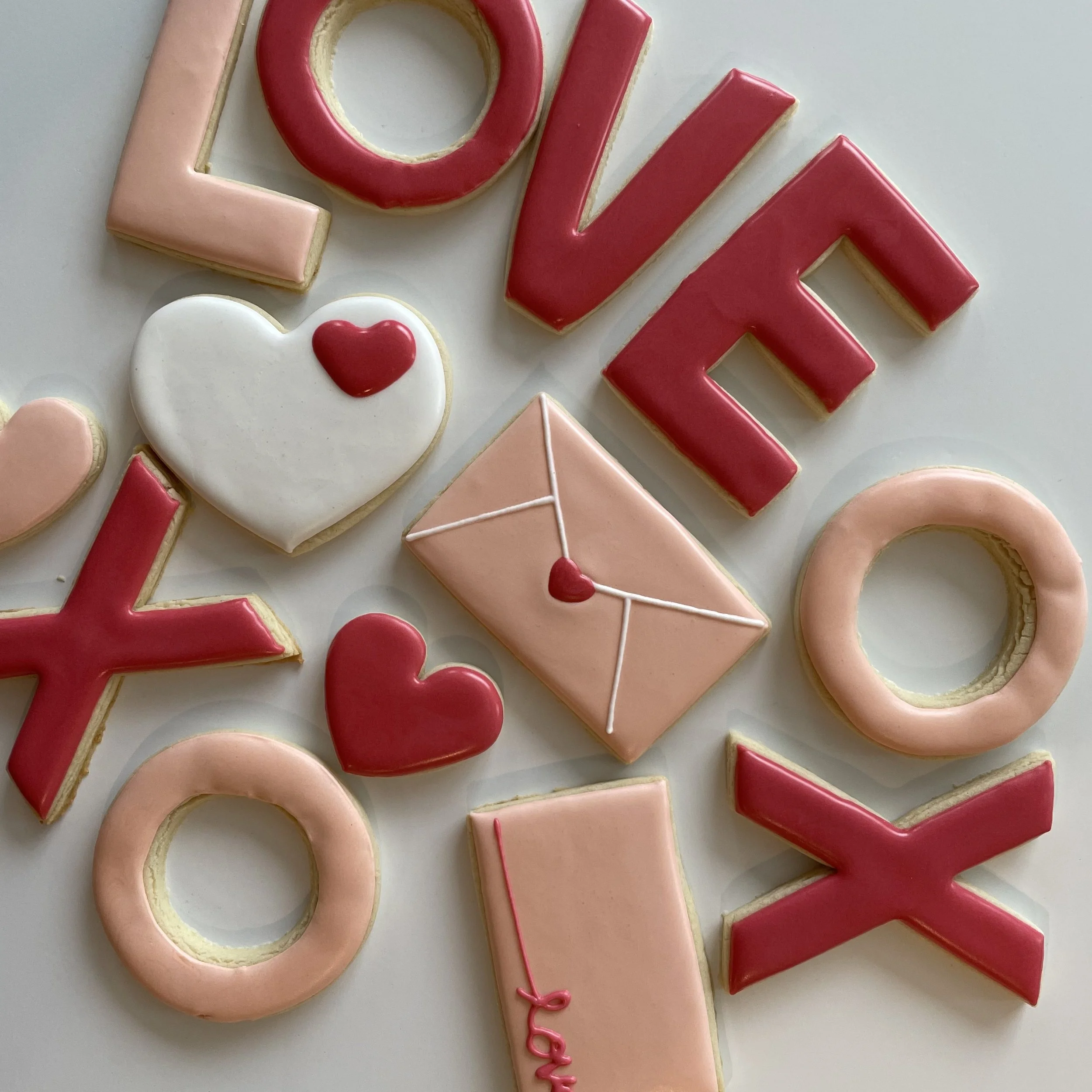 Decorative cookies spelling out the word "LOVE" in pink and red icing, with a heart-shaped cookie featuring a smaller red heart on white icing, on a white background.