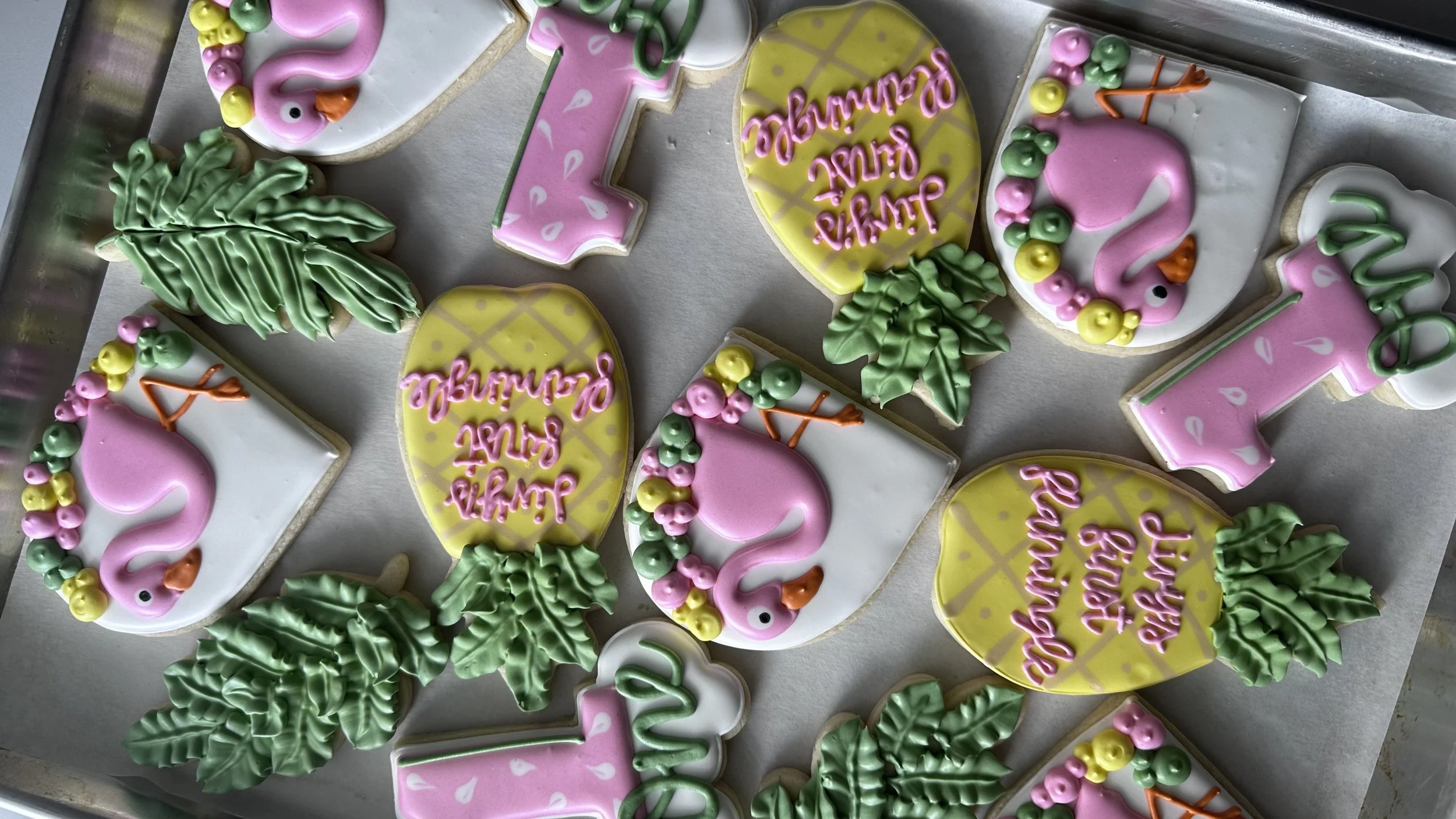 Decorated cookies with pink flamingo designs, green palm trees, and yellow pineapples, with some cookies featuring the phrase 'Enjoy the Sunshine.'