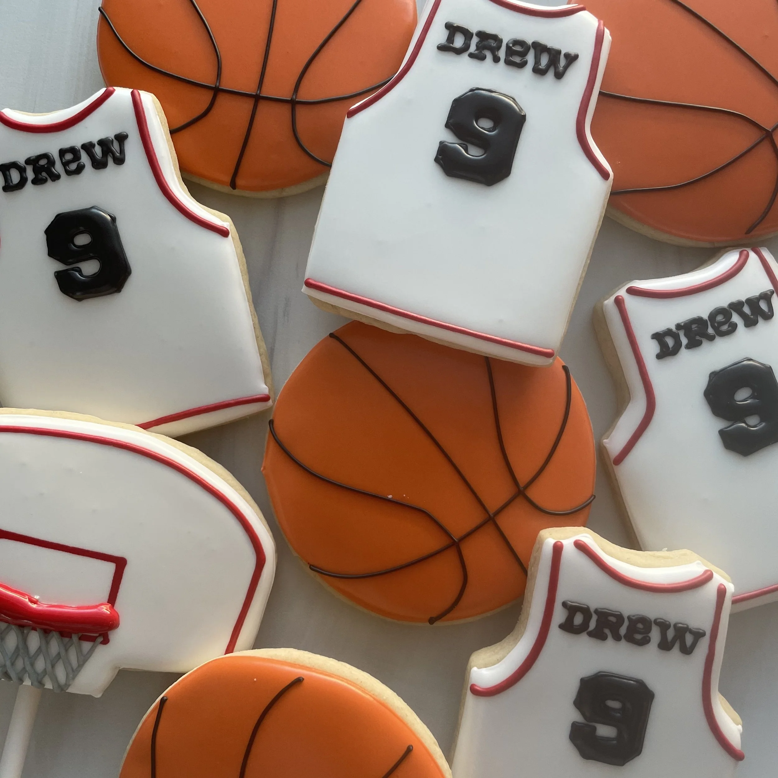 Cookies decorated with white, orange, and black icing in basketball and sports theme, with text 'Drew 9' on some cookies, set against a background of orange and black basketball shapes.