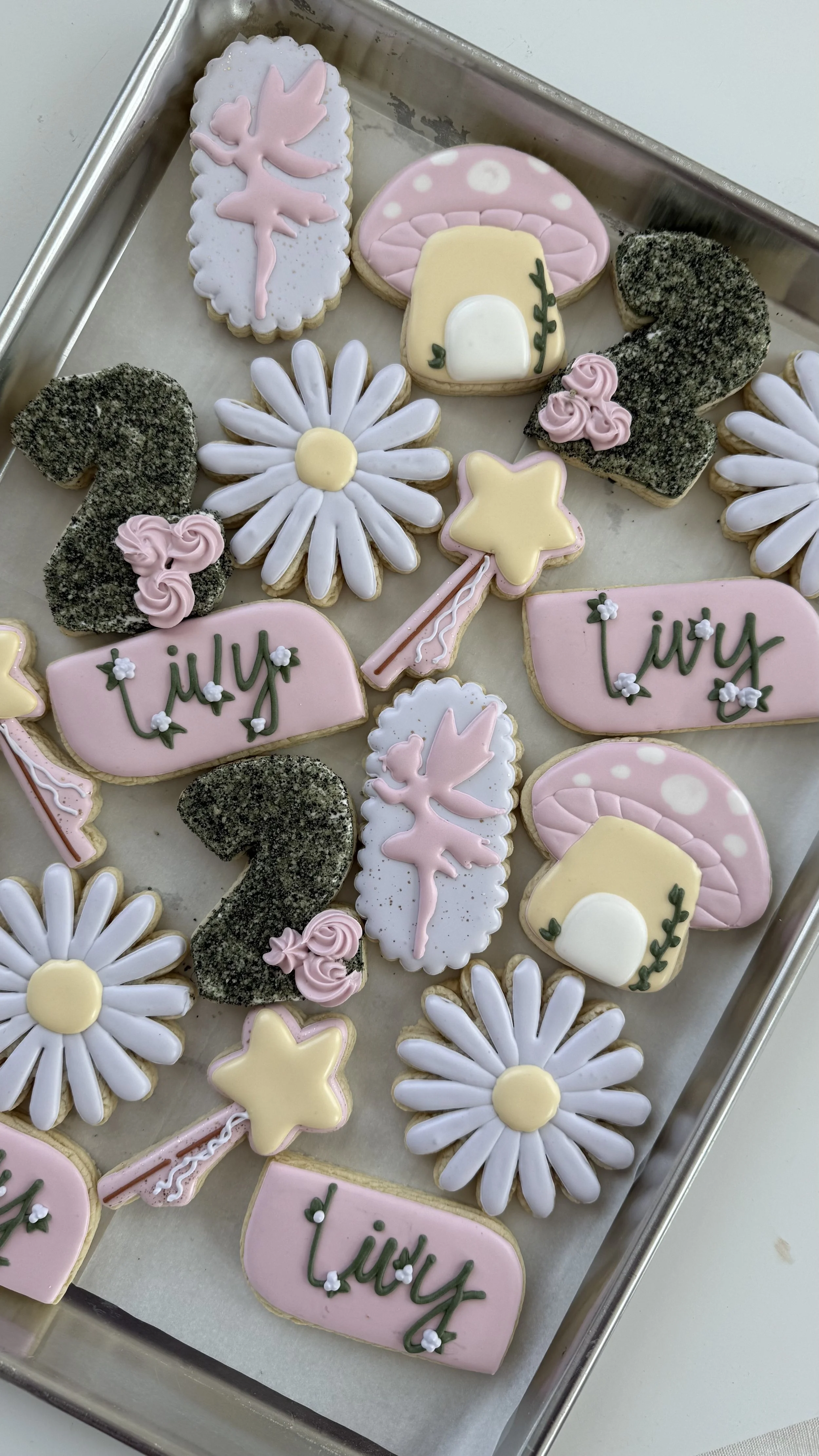 Assorted decorated cookies with summer themes, including daisy flowers, mushrooms, pink stars, black sugar-coated numbers, and pink banners with the word 'lily' written in green, arranged on a silver tray.