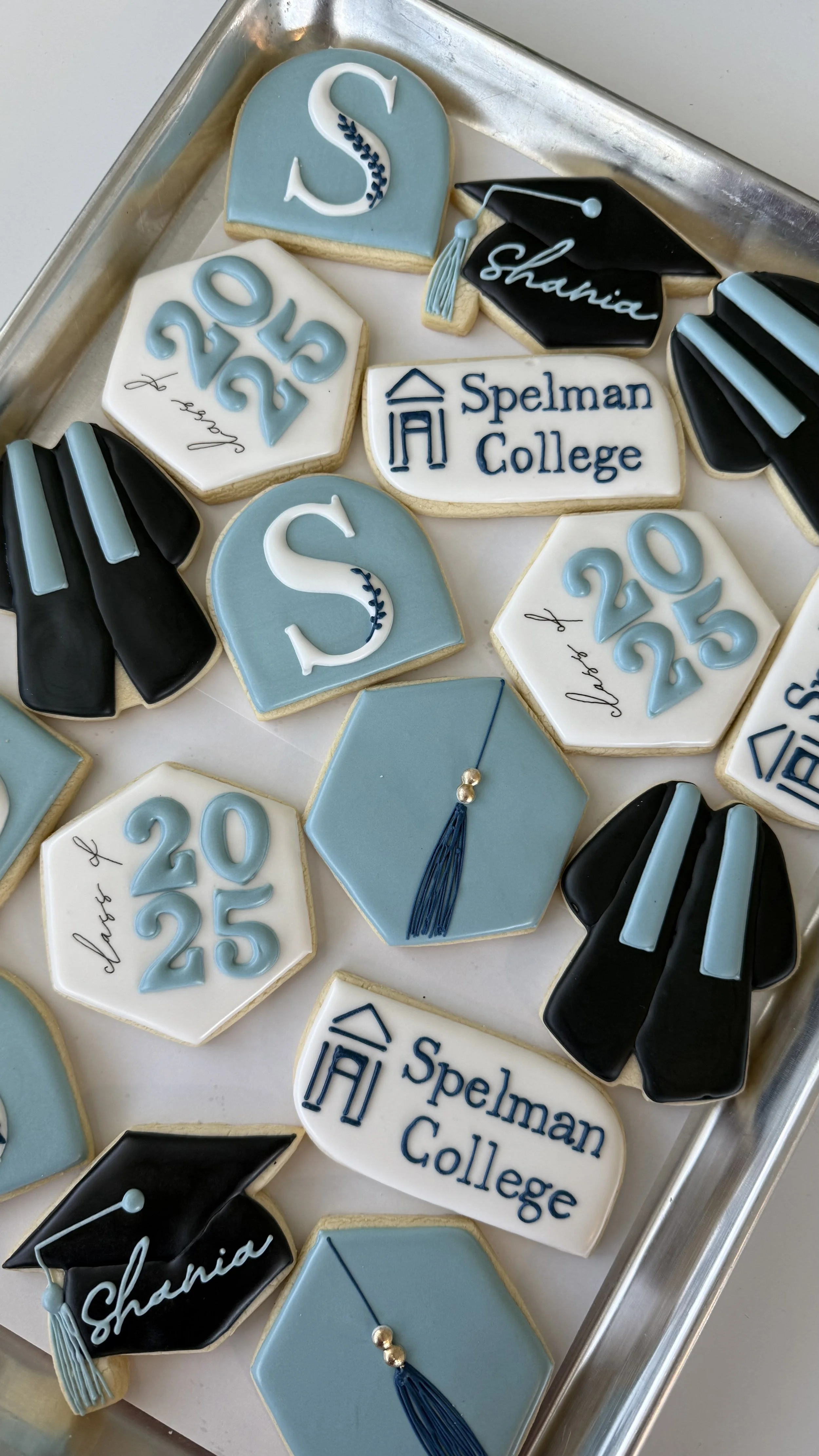 Decorated cookies celebrating a graduation with academic motifs, including diplomas, mortarboards, and personalized names and dates in blue, black, and white colors.
