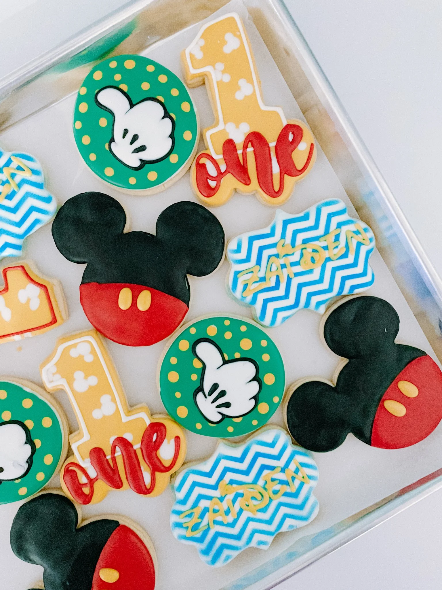 Decorated cookies with Disney-themed designs including Mickey Mouse ears, a thumbs-up symbol, the number one, and the word "love" in red lettering, arranged in a clear container.