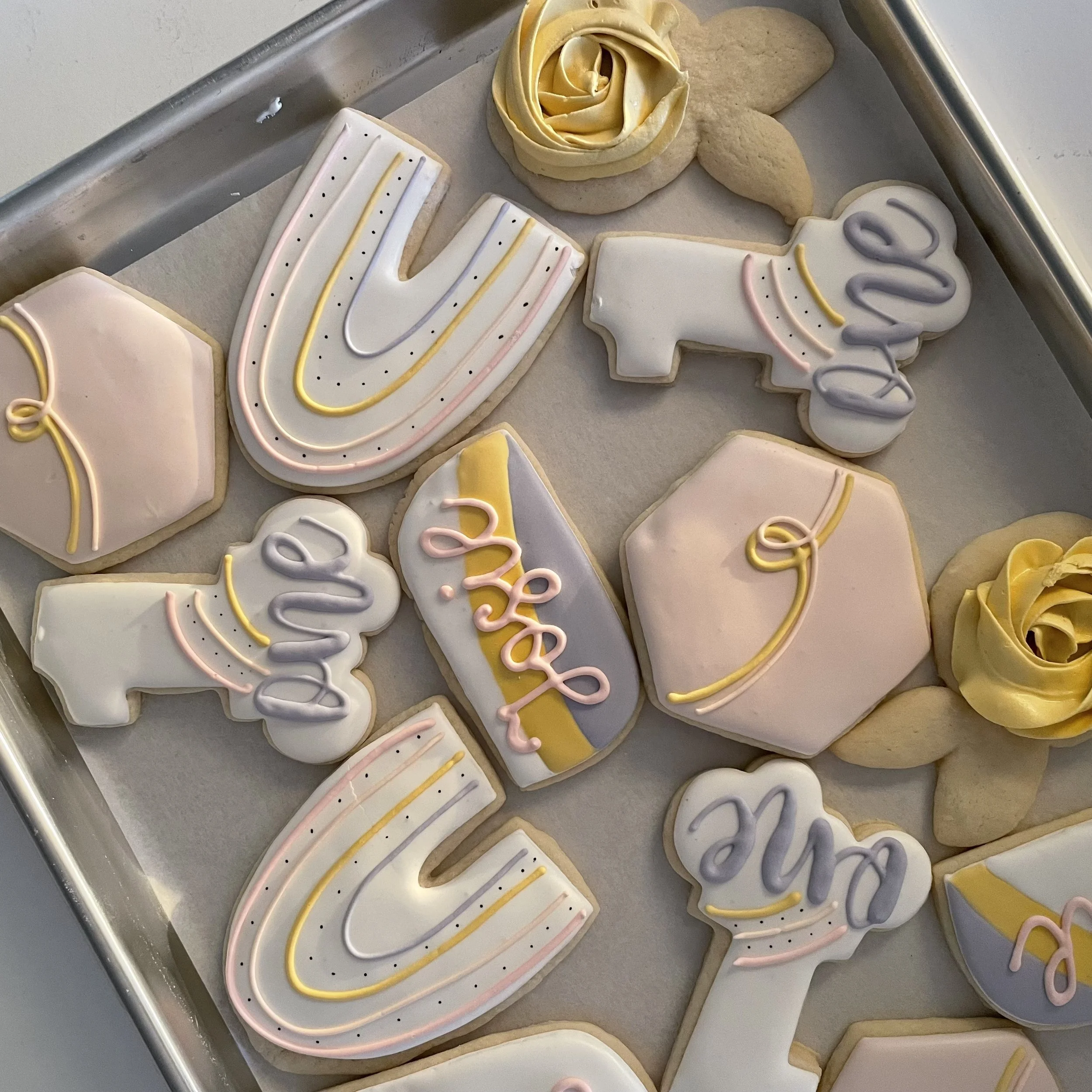 Assorted decorated sugar cookies on a baking sheet, featuring letters, numbers, and festive shapes with pastel and bright icing decorations.