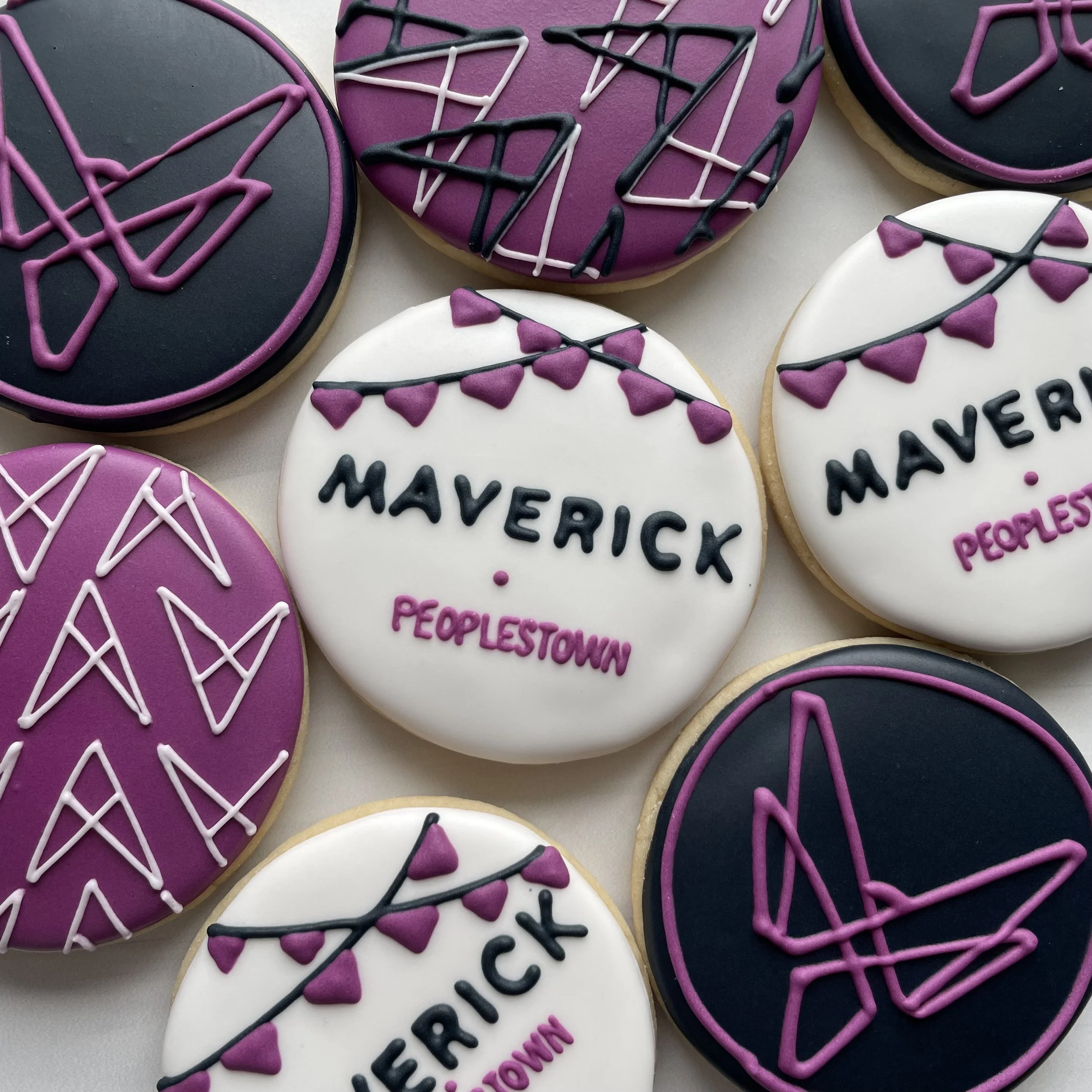 Decorated cookies with purple, black, and white icing designs, some with hearts and text, on a light background.