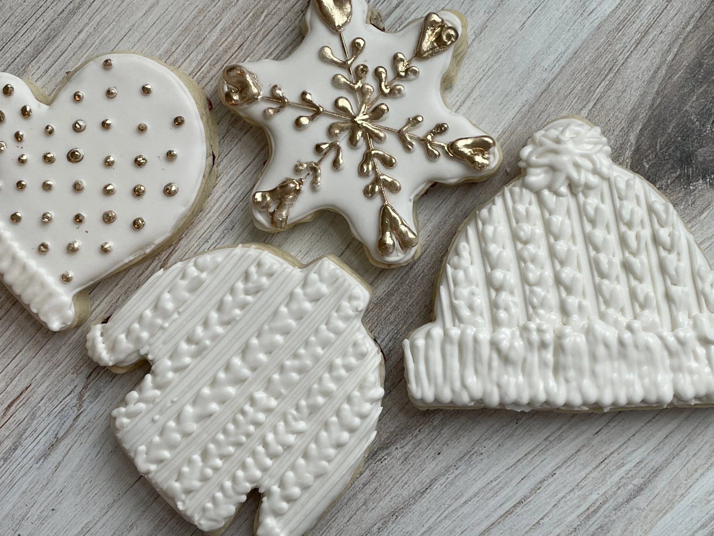 Assorted decorated cookies on a wooden surface, including a snowflake-shaped cookie with gold accents, heart-shaped cookies with white icing and small gold dots, and a Christmas tree-shaped cookie with white icing and pearl-like decorations.