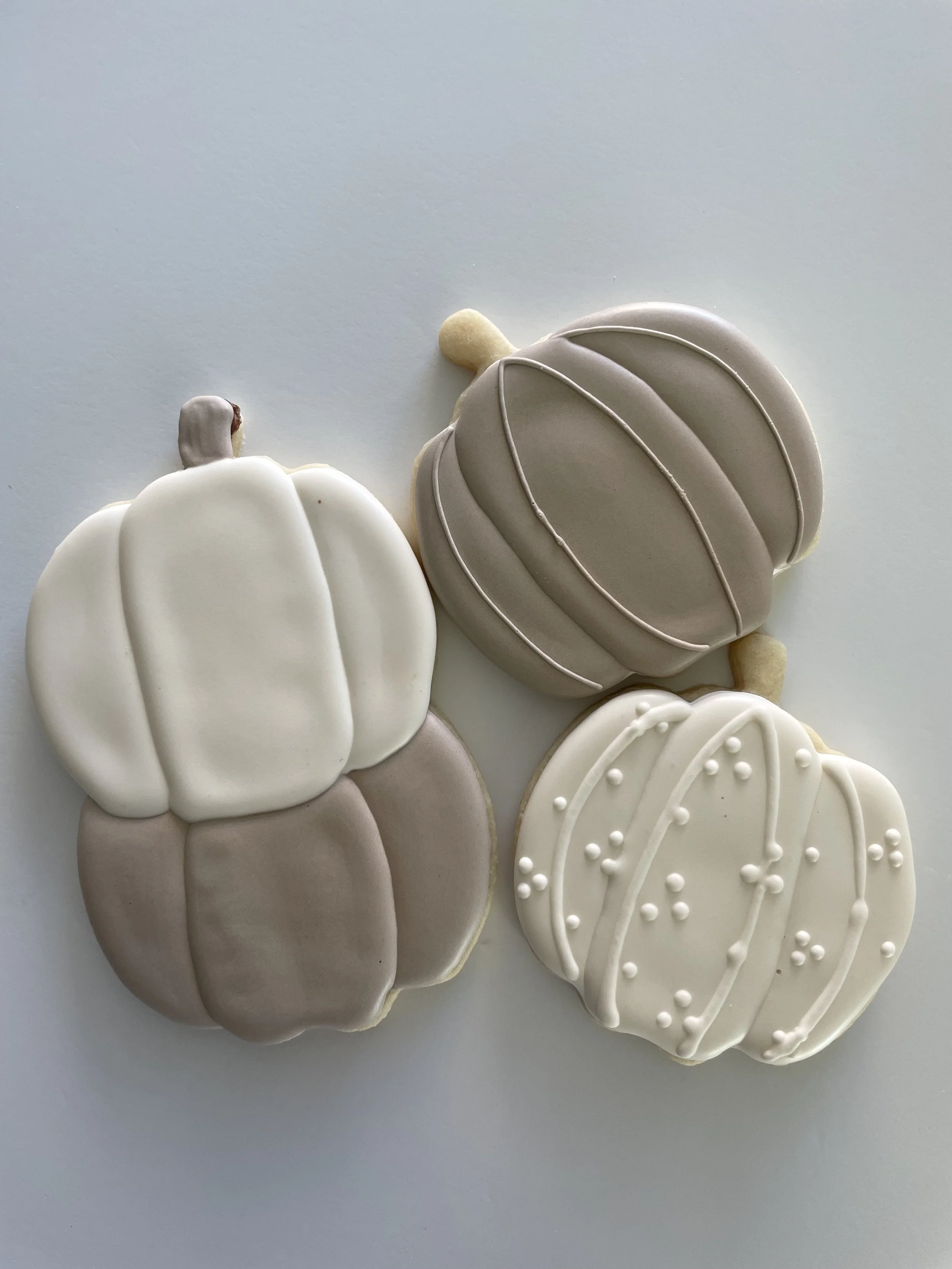 Decorated sugar cookies shaped like pumpkins, with white, taupe, and beige icing, some with piped lines and dots, on a white surface.