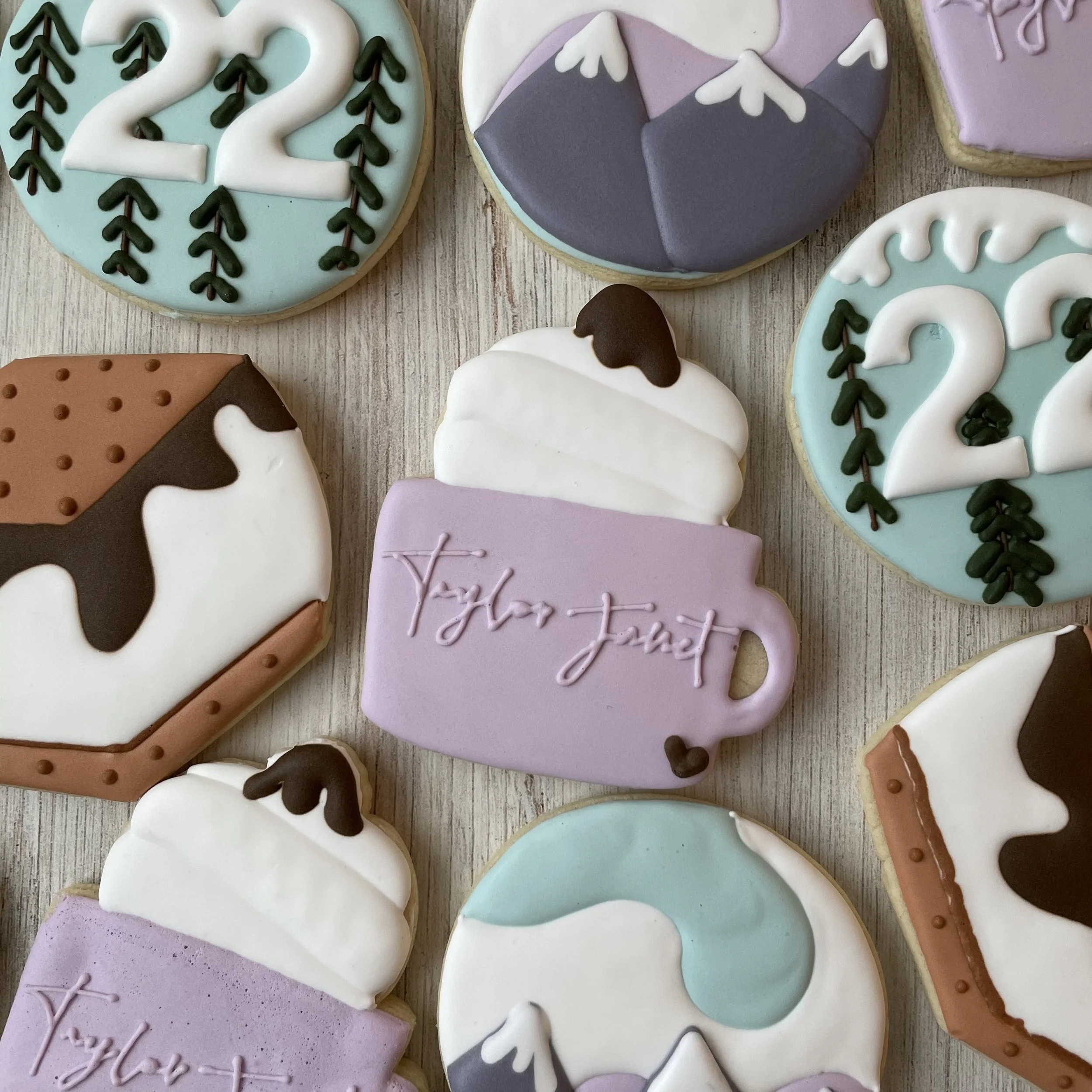 Decorative sugar cookies with pastel purple, white, gray, blue, and brown icing in various shapes, including hearts, snowflakes, and a mountain, some with personalized messages written in icing.