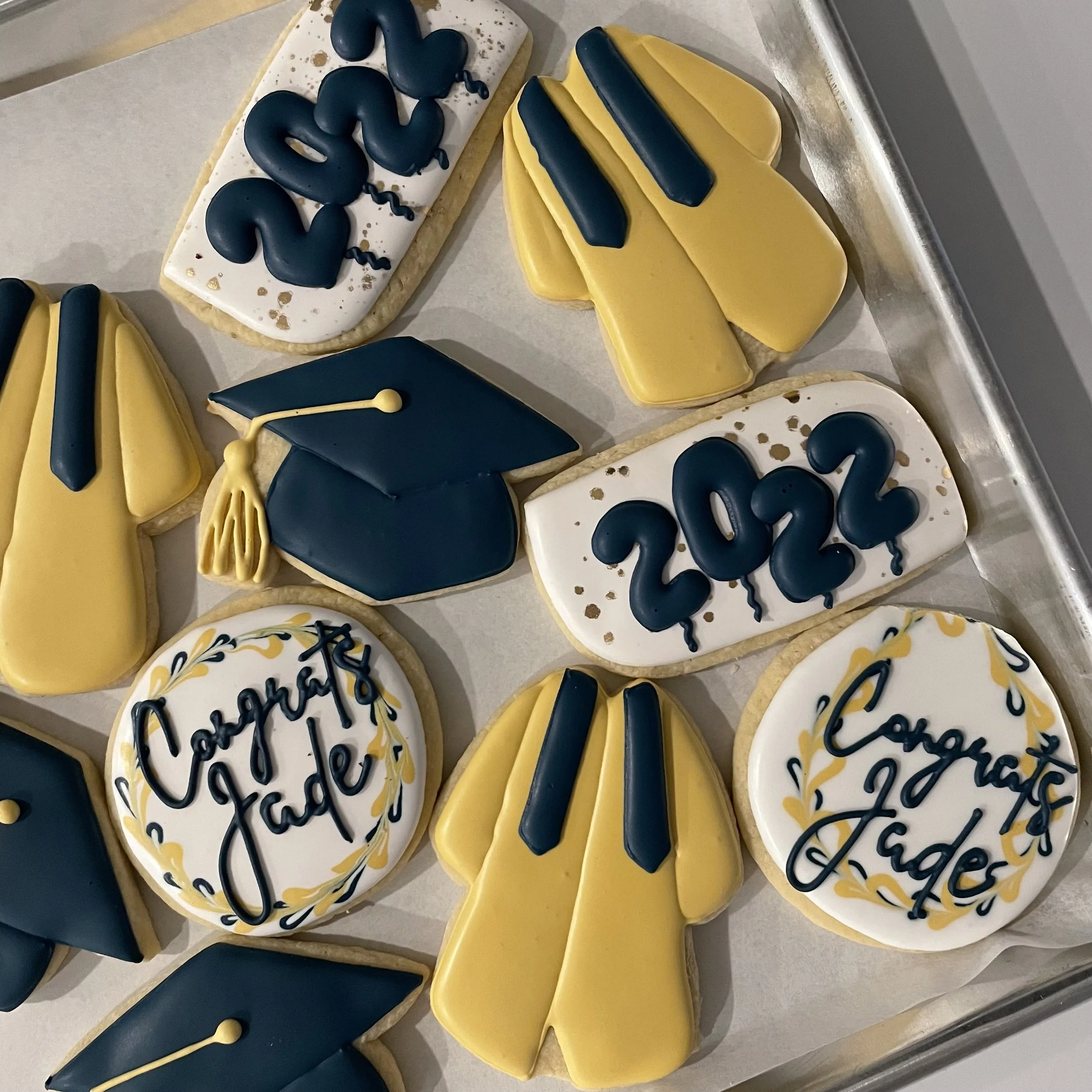 Decorated cookies with yellow, white, and dark blue icing, some with written words, arranged on a tray.