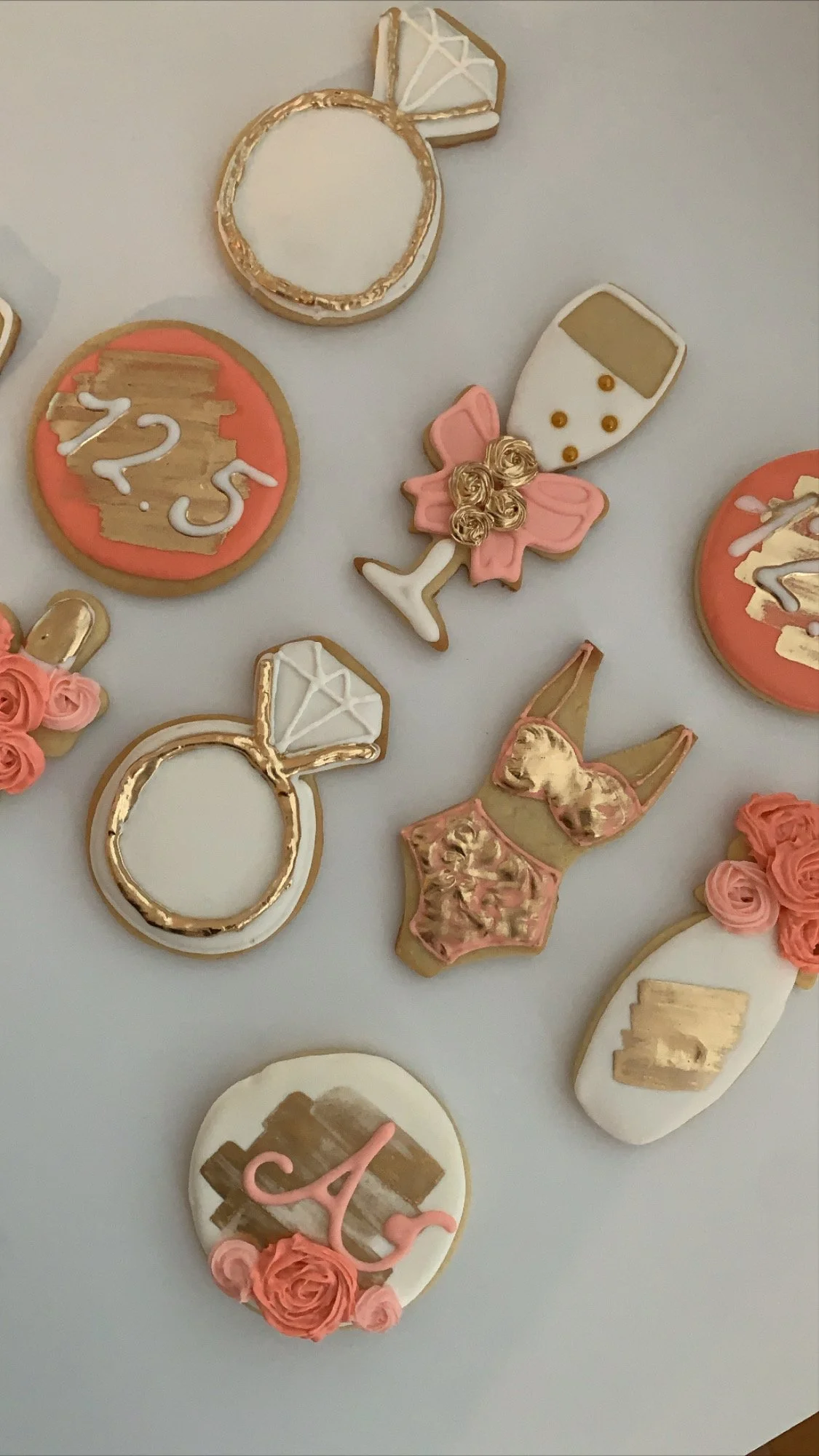 Decorative cookies shaped like a diamond ring, champagne glass, roses, and the number 23, with pink and gold accents, on a white surface.