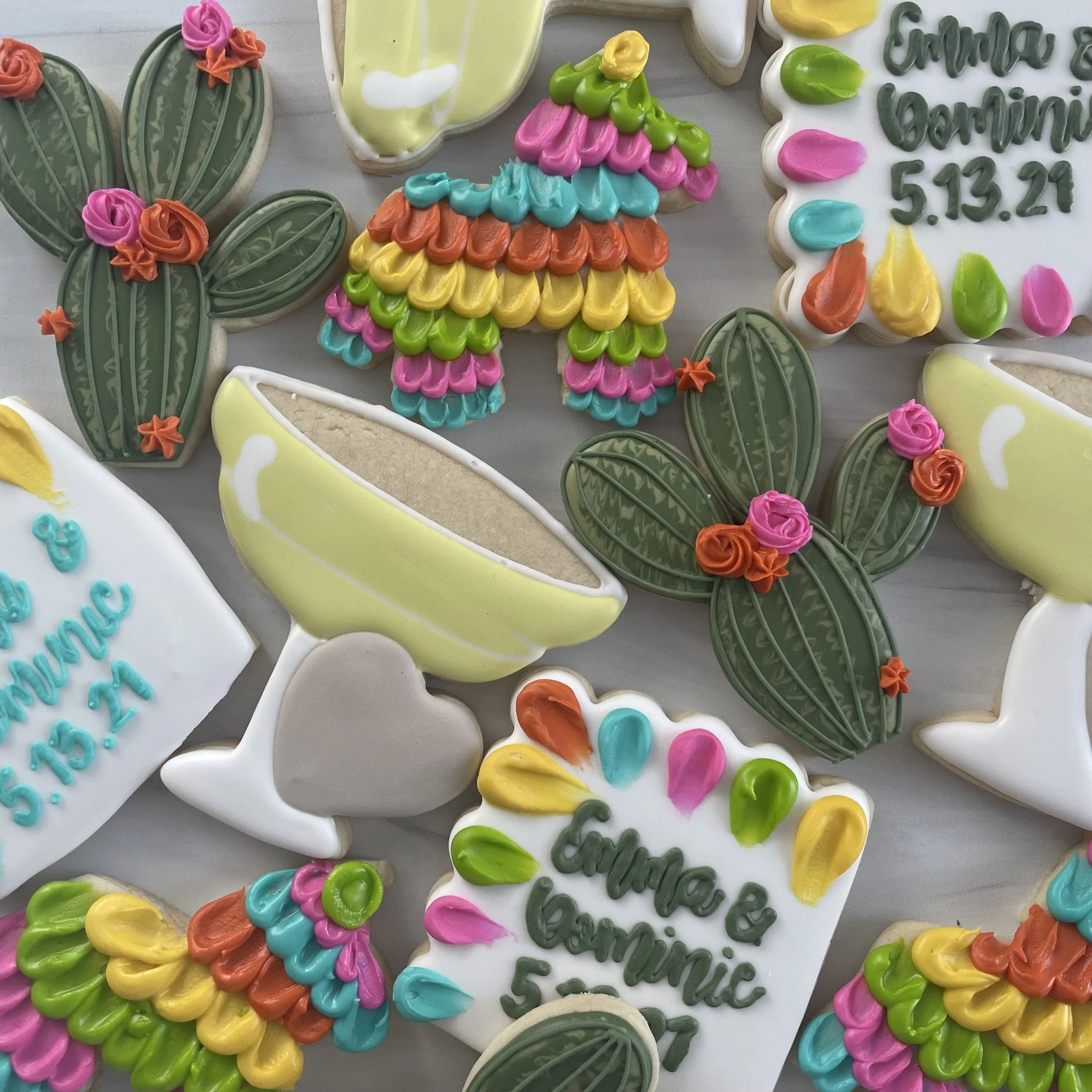 Colorful decorated cookies in various shapes including cactus, rainbow, and cactus with flowers, with some cookies containing written messages or dates.