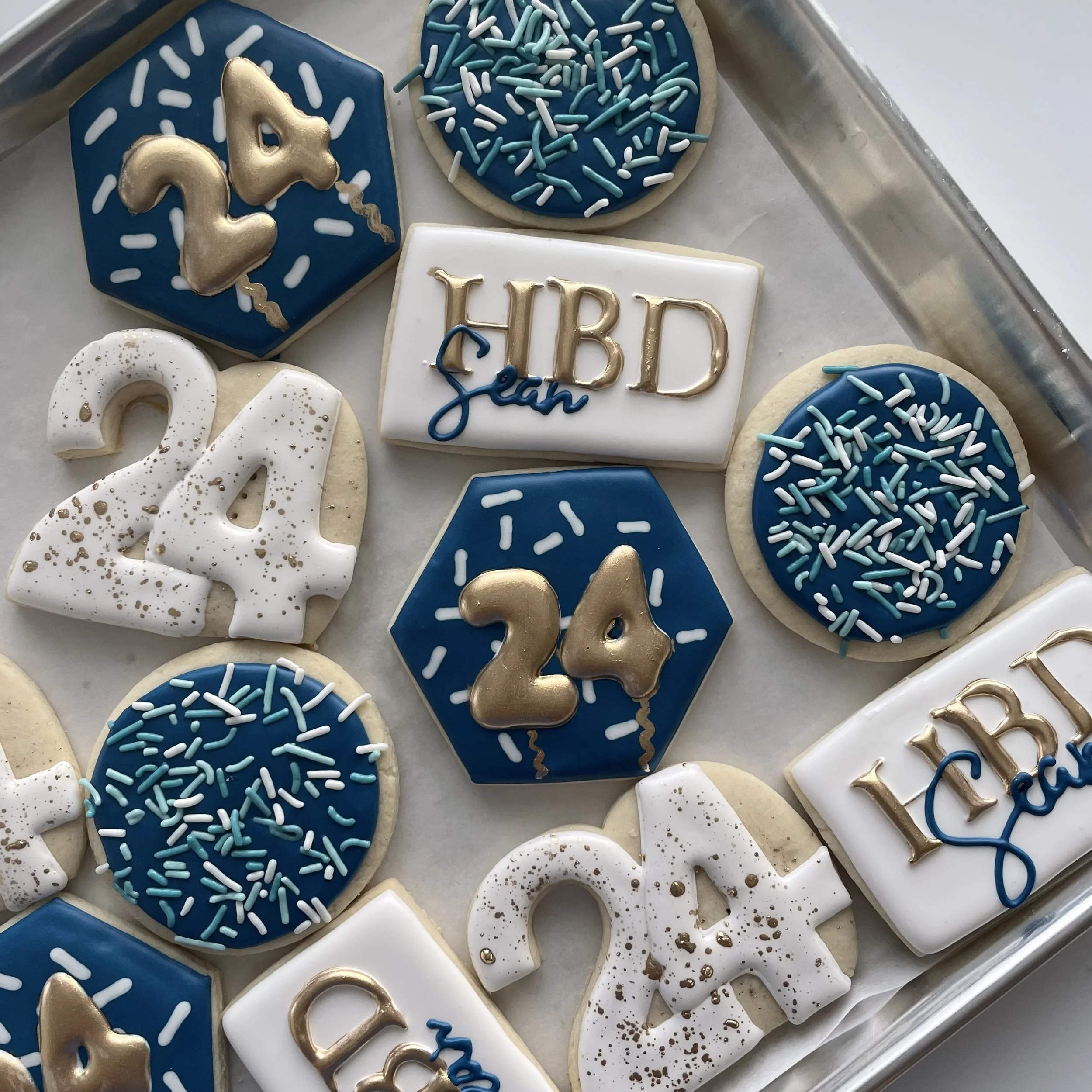 Cookies decorated for a celebration, including a blue hexagon cookie with a gold unicorn candle, round cookies with blue icing and sprinkles, and a white rectangular cookie with gold lettering that says 
