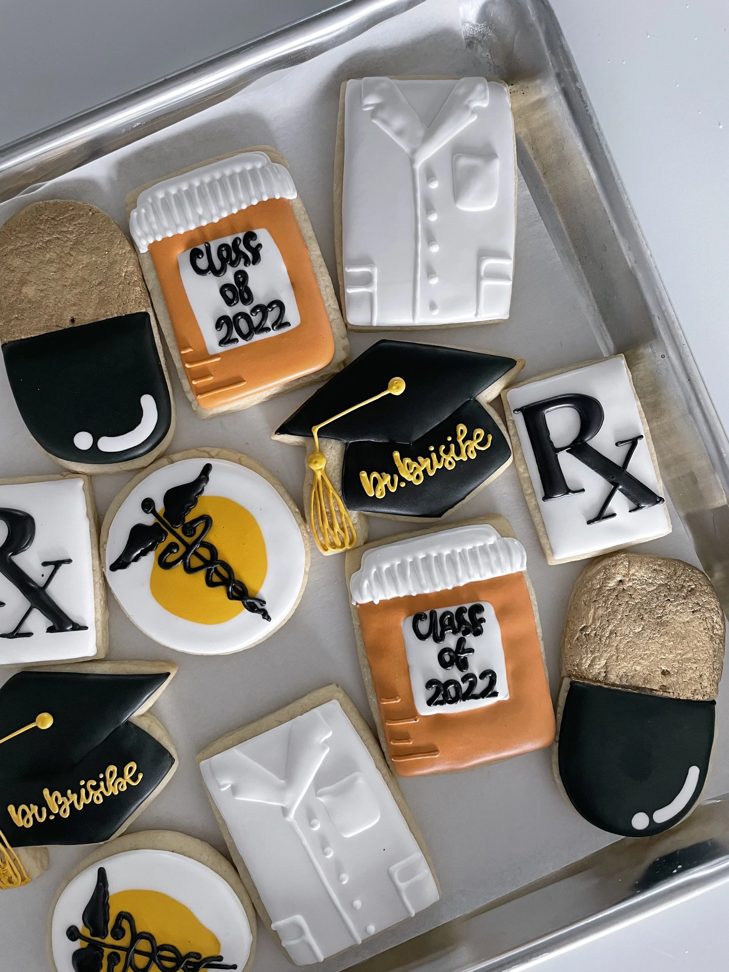 Assorted school-themed cookies decorated with icing, including a graduation cap, diploma, and a computer monitor, on a baking sheet.