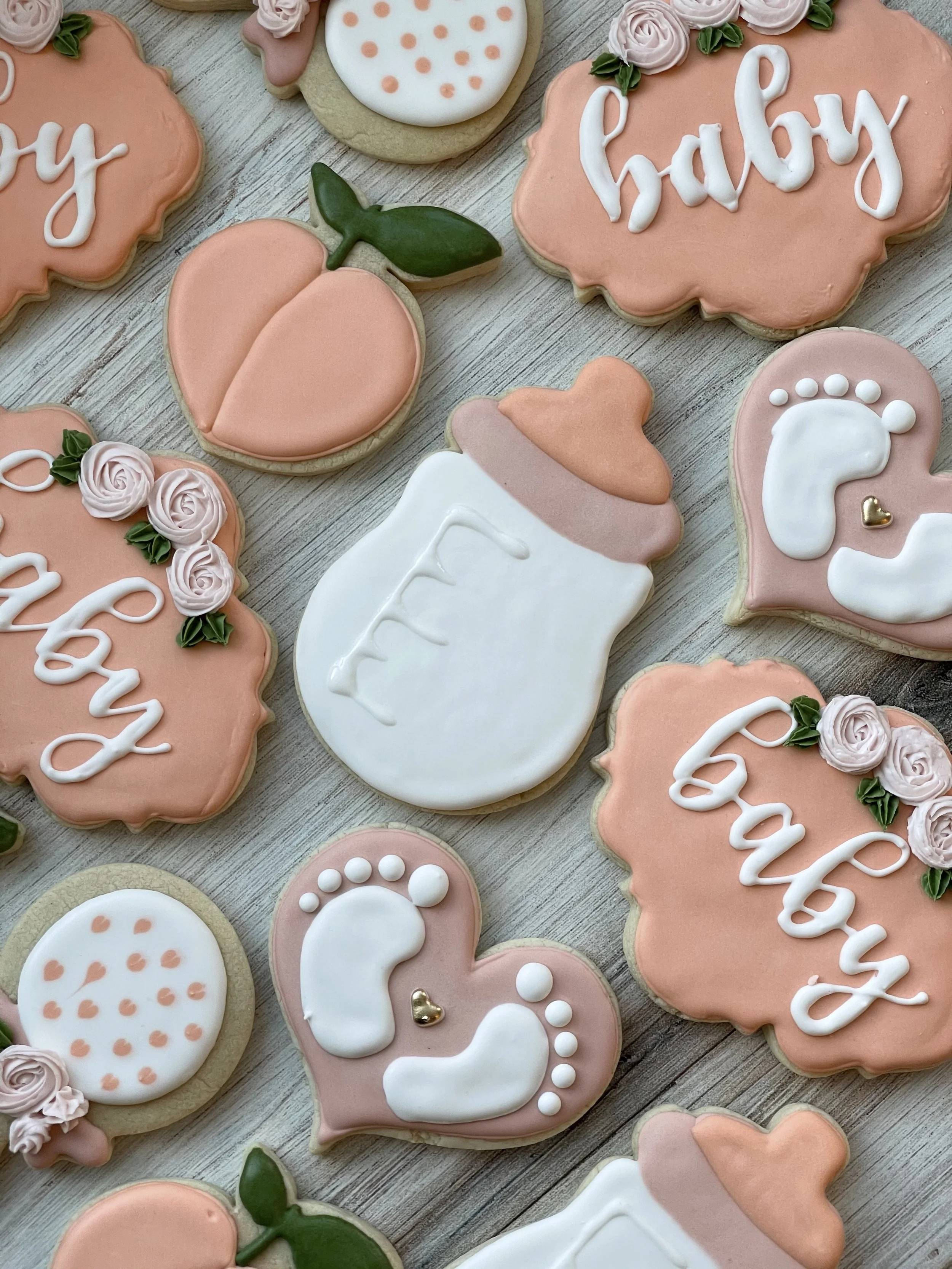 Assorted decorated sugar cookies with pink, white, and beige icing, some with floral and leaf designs, and others with the word 'baby' written in white icing, arranged on a wooden surface.