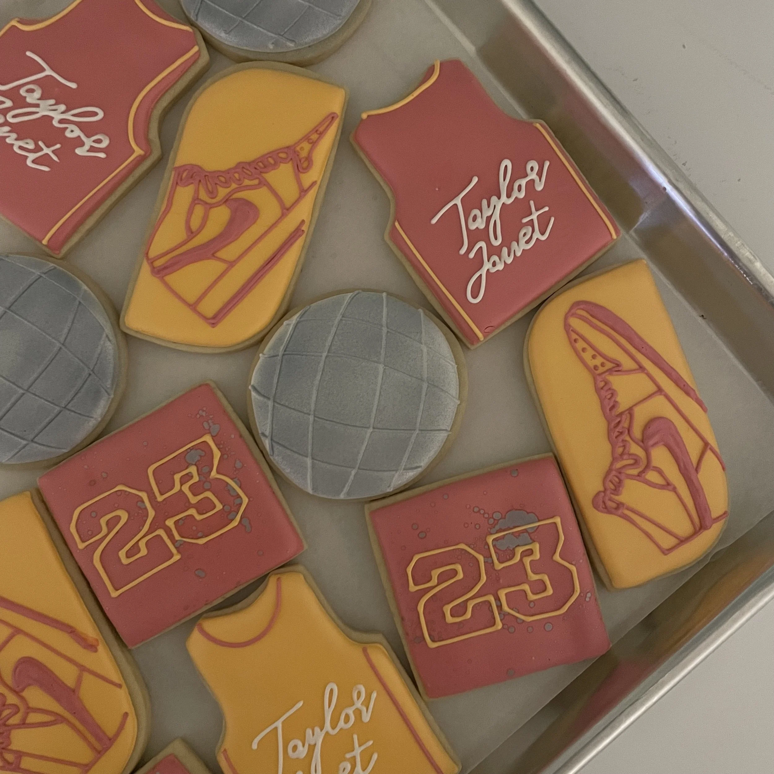 Close-up of decorated cookies with pink and yellow icing, some with white text, on a silver baking sheet.