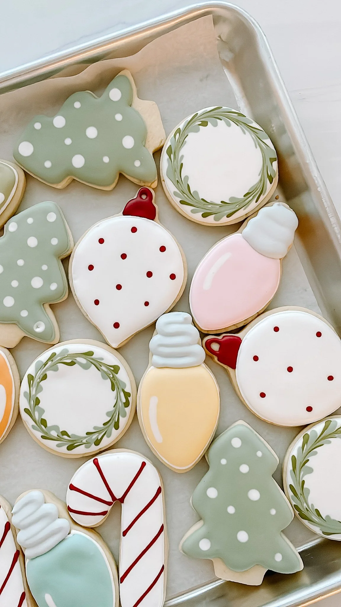 Assorted holiday cookies decorated with pastel icing, including Christmas trees, ornaments, candy canes, and wreaths on a baking sheet.
