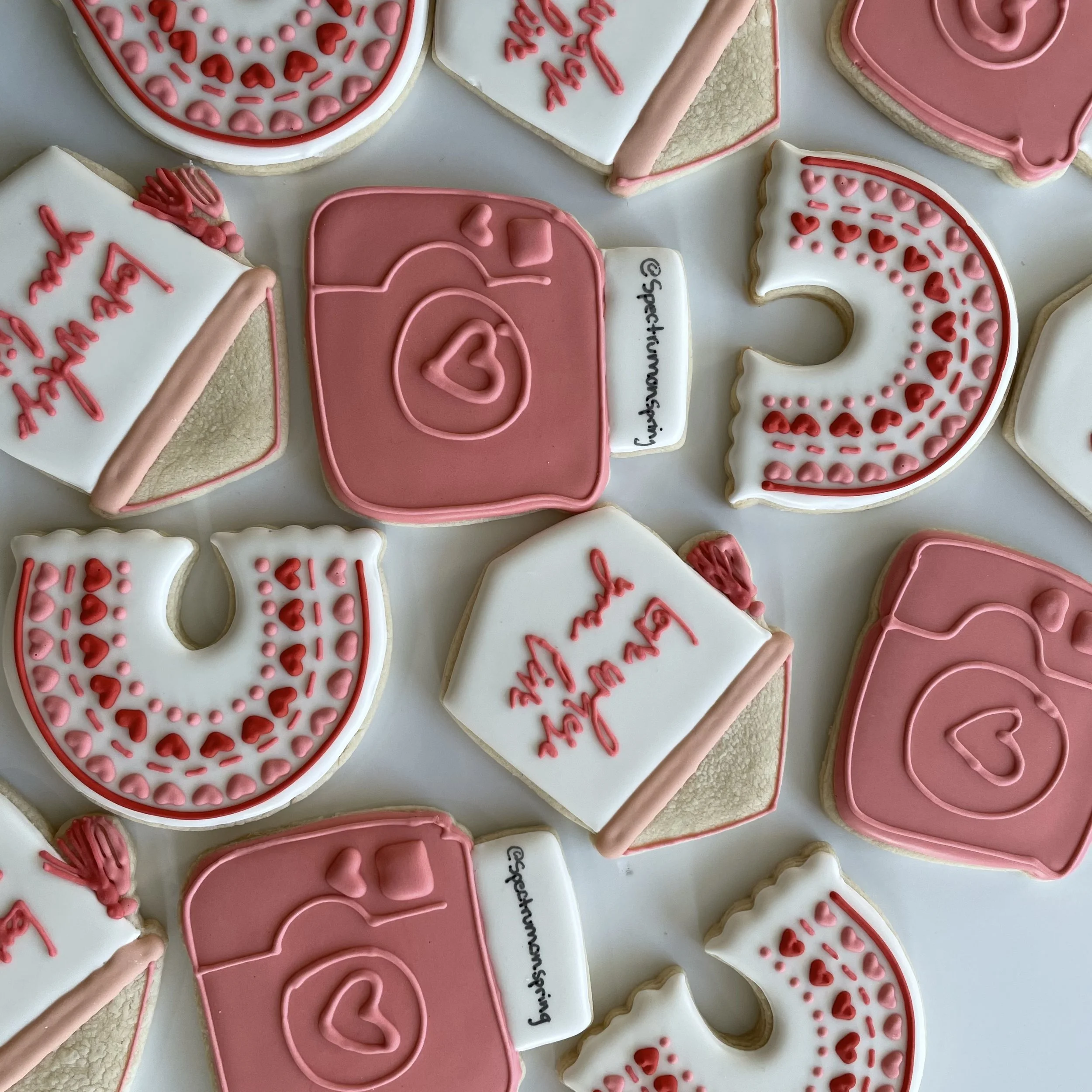 Decorative cookies with pink and white icing, some with heart patterns, text, and pink ribbons, arranged on a flat surface.