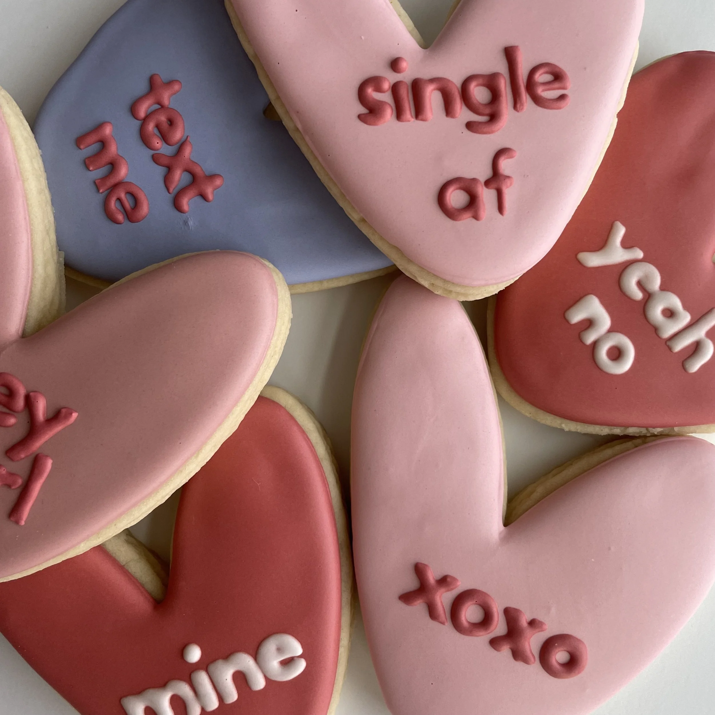 Close-up of heart-shaped cookies decorated with pink and purple icing. Some have the word 'love' written in pink icing and others have the phrase 'be happy' in pink icing. The cookies are arranged overlapping each other on a light-colored surface.