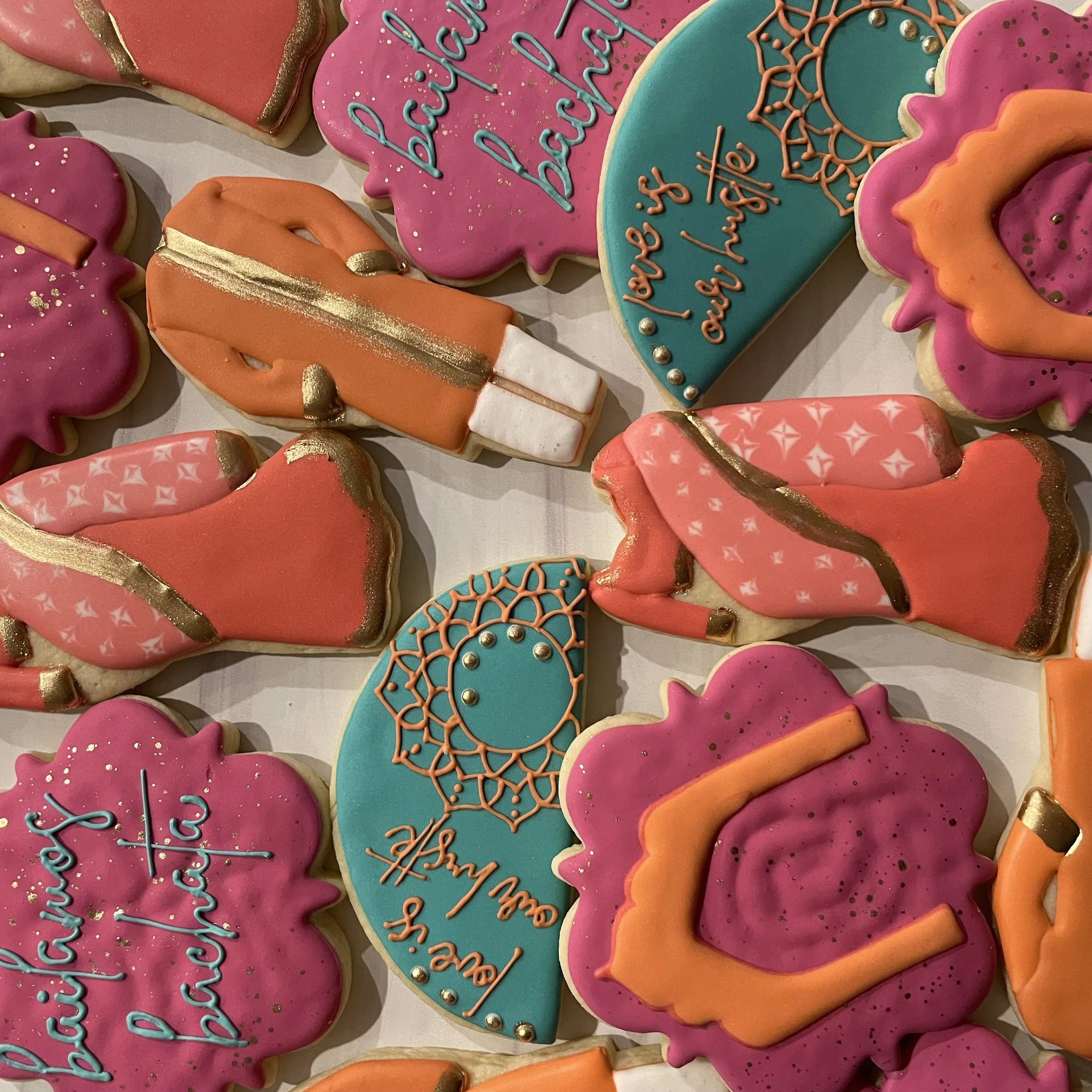 Assorted decorated cookies for a celebration, featuring pink, blue, and orange icing with messages such as "Congratulations" and "Best Wishes" alongside decorative embellishments like gold accents, sprinkles, and intricate icing designs.
