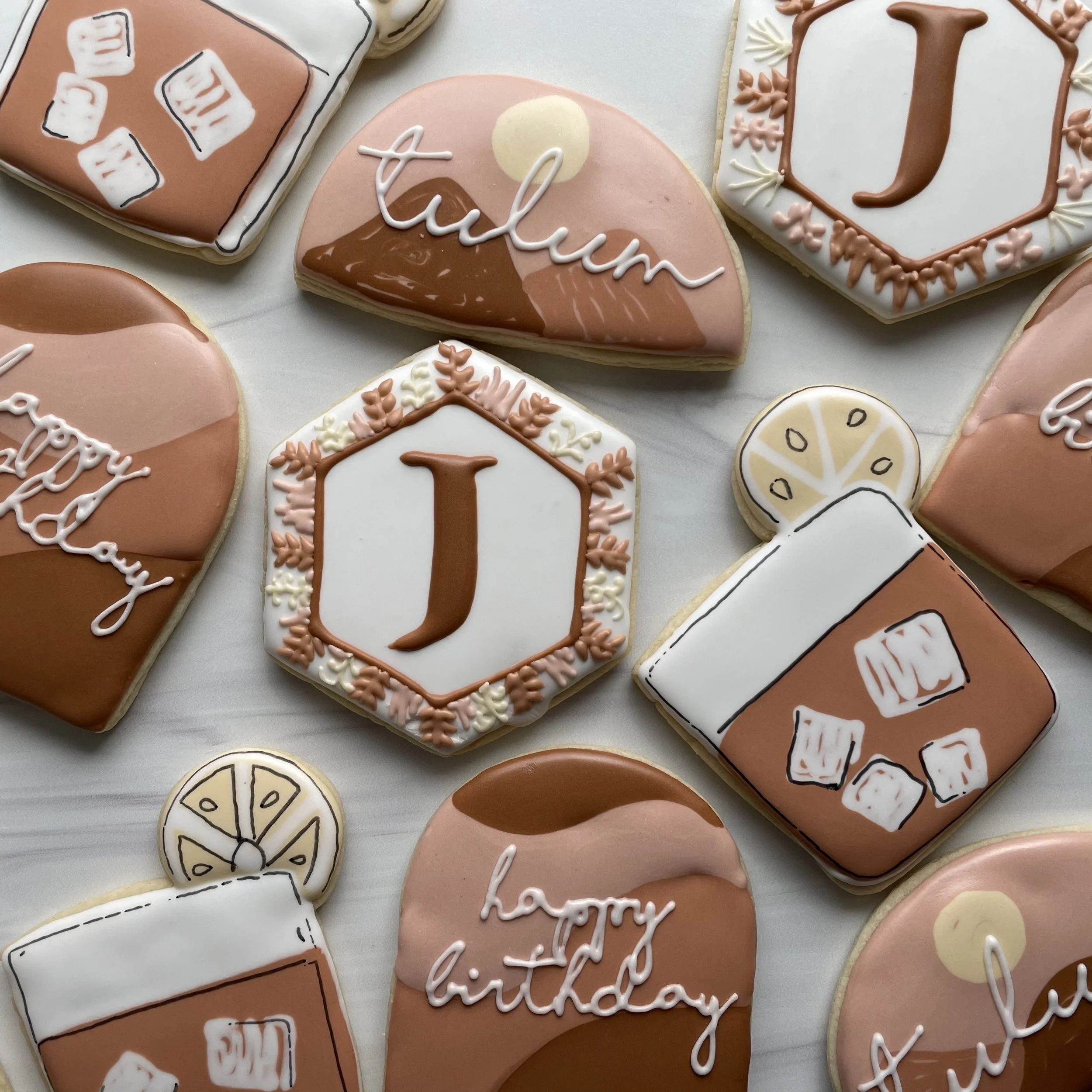 Assorted decorated cookies for a celebration, featuring pink, white, and brown icing with messages like 'happy birthday' and 'thank you,' and designs such as geometric patterns, flowers, and text.