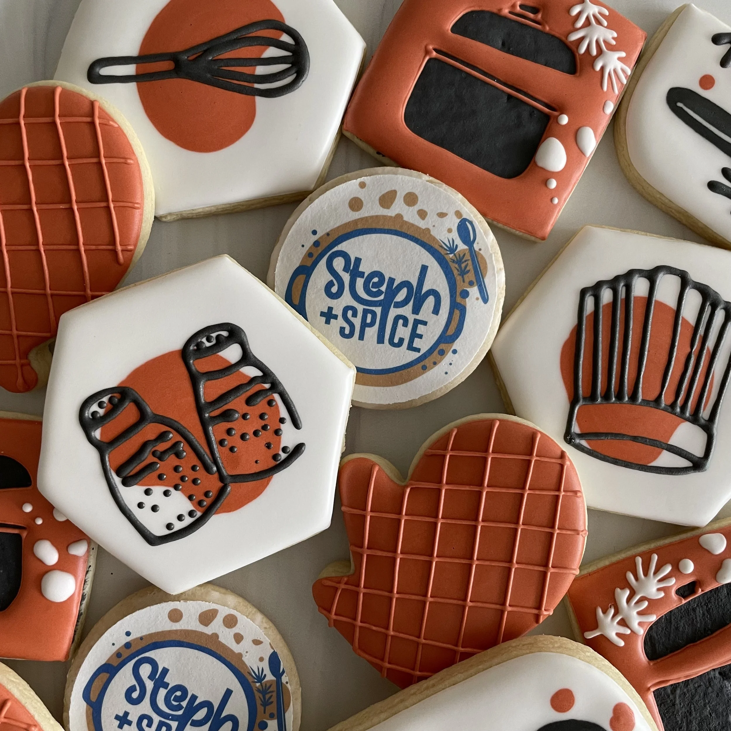 Decorated sugar cookies with holiday or winter theme, including cookies with snowflake patterns, a cookie with a cooking pot, and others with black, white, red, orange, and blue icing details.