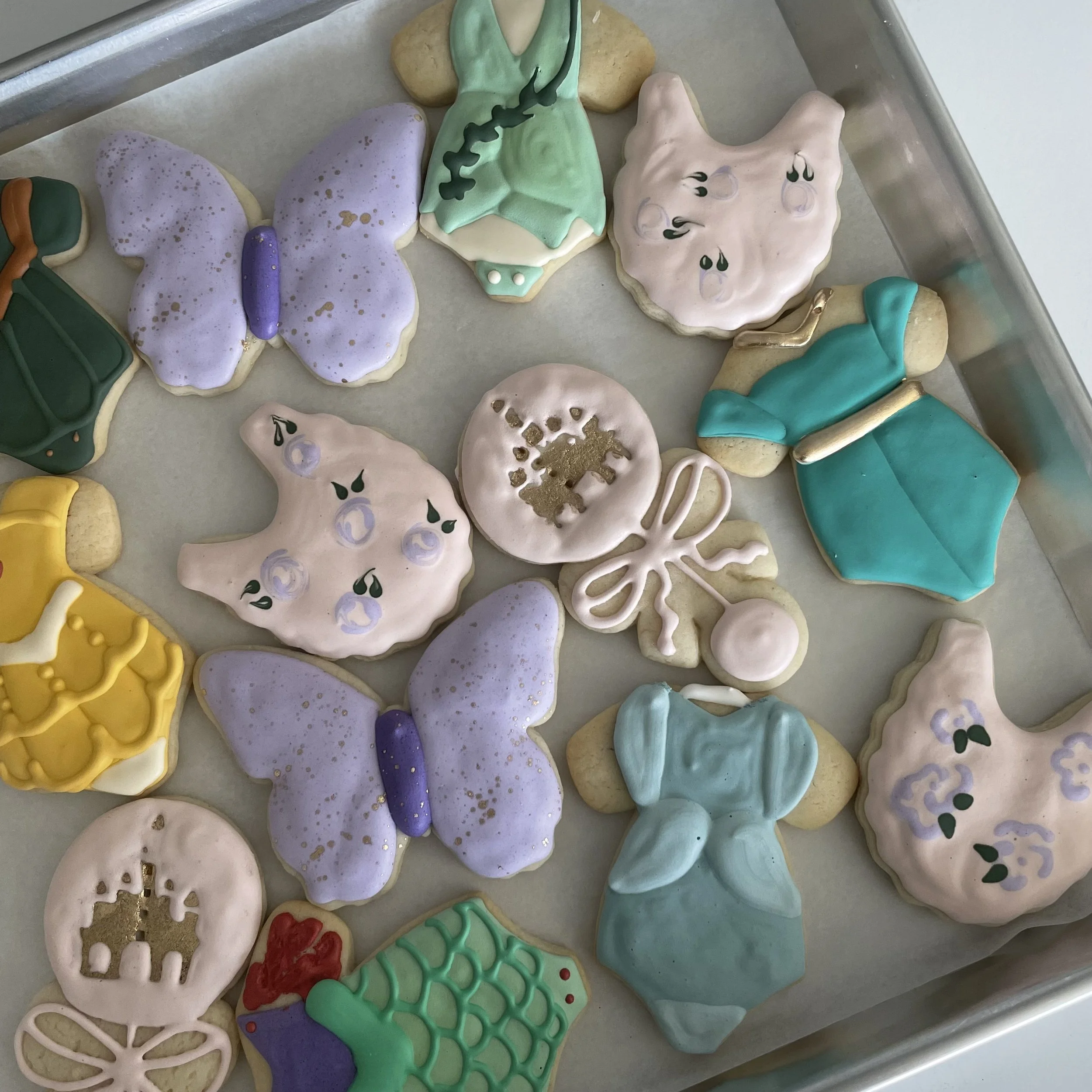 Assorted decorated cookies on a baking sheet, featuring pastel colors, floral, butterfly, and abstract patterns.