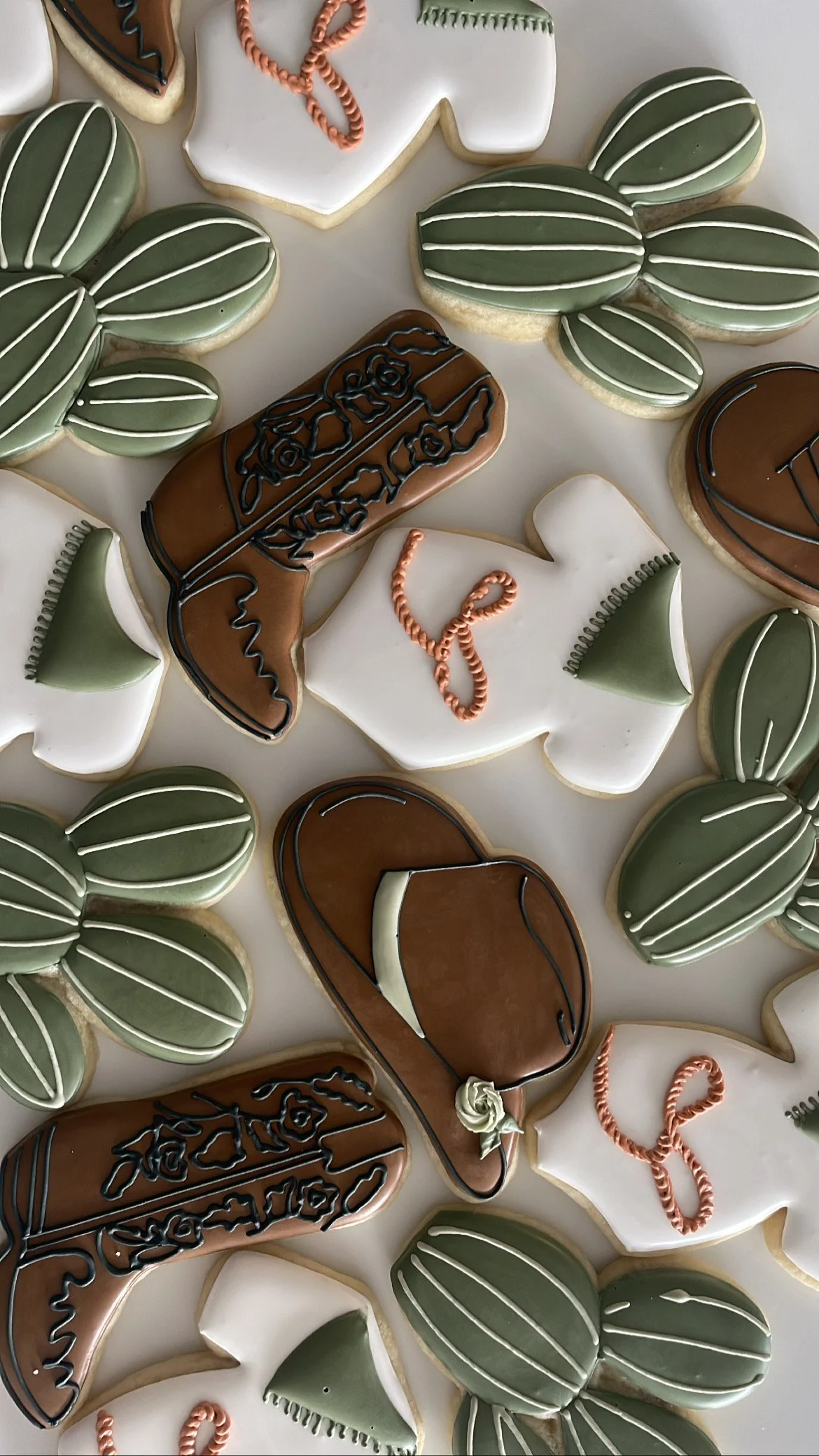 Decorative cookies shaped like cacti, leather boots, and desert accessories, decorated with icing in green, brown, white, and black colors.