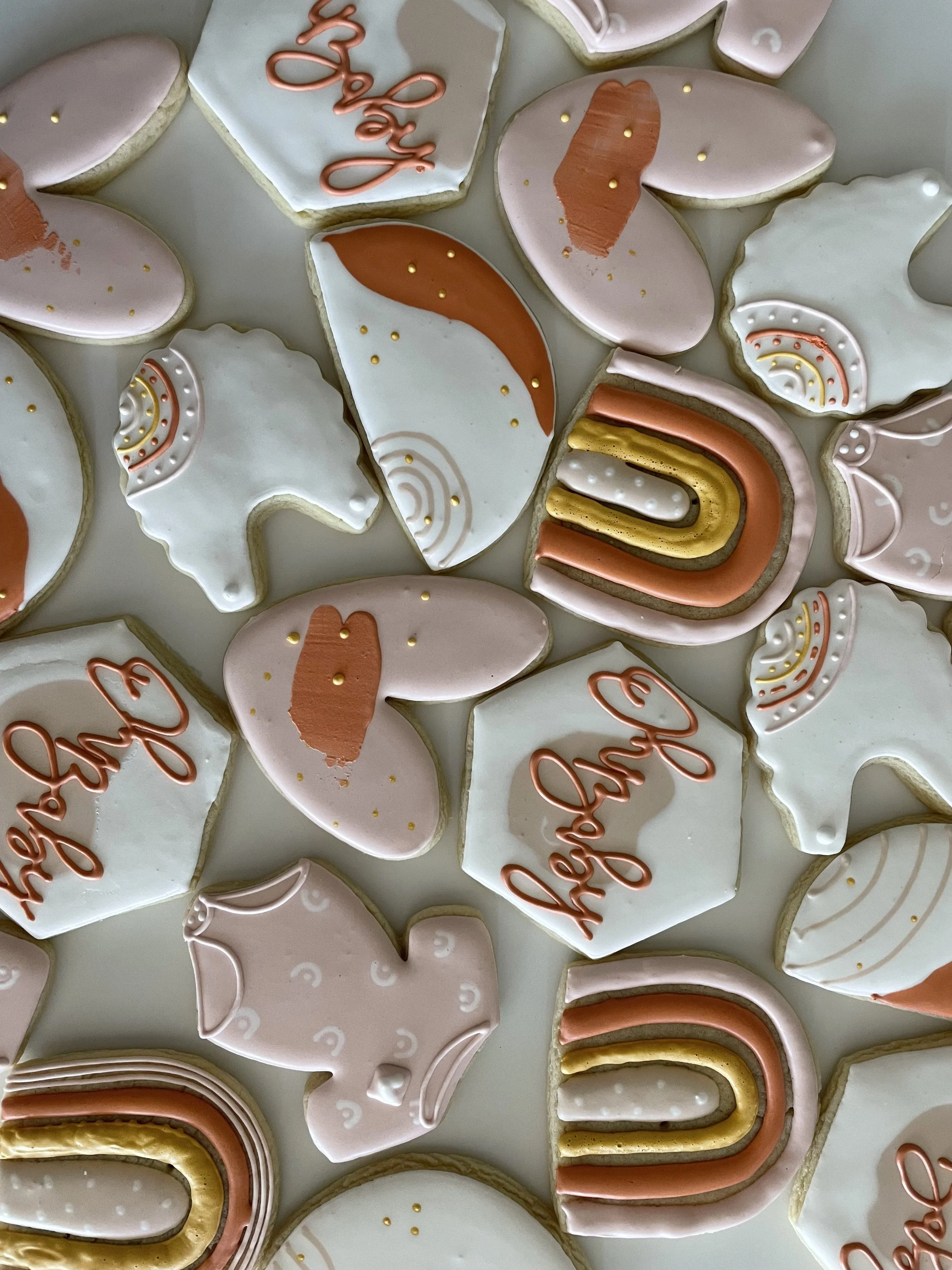Close-up of decorated cookies with pink, white, and gold icing, featuring rainbow, heart, and abstract designs, with some cookies displaying the word 'Love' in pink icing.