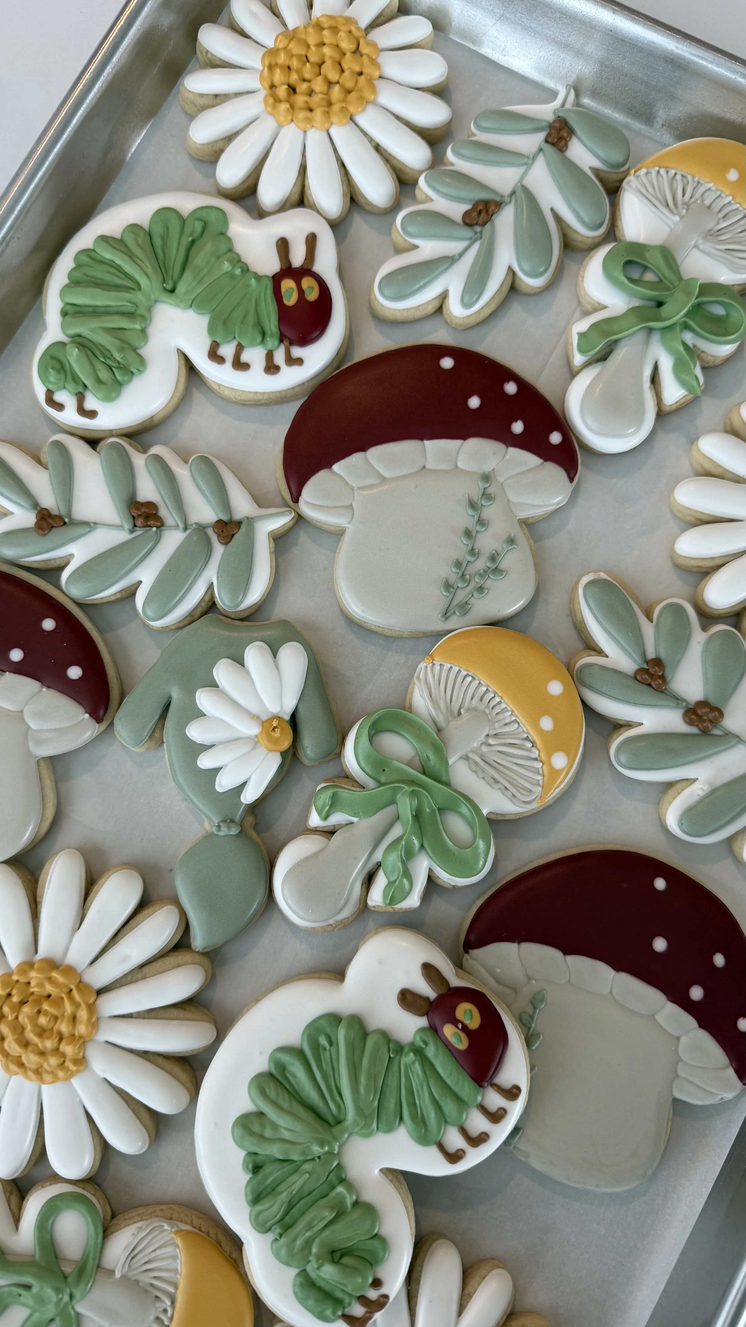 Decorative cookies with floral and nature-inspired designs, including daisies, mushrooms, leaves, and a caterpillar with a red head and green body.