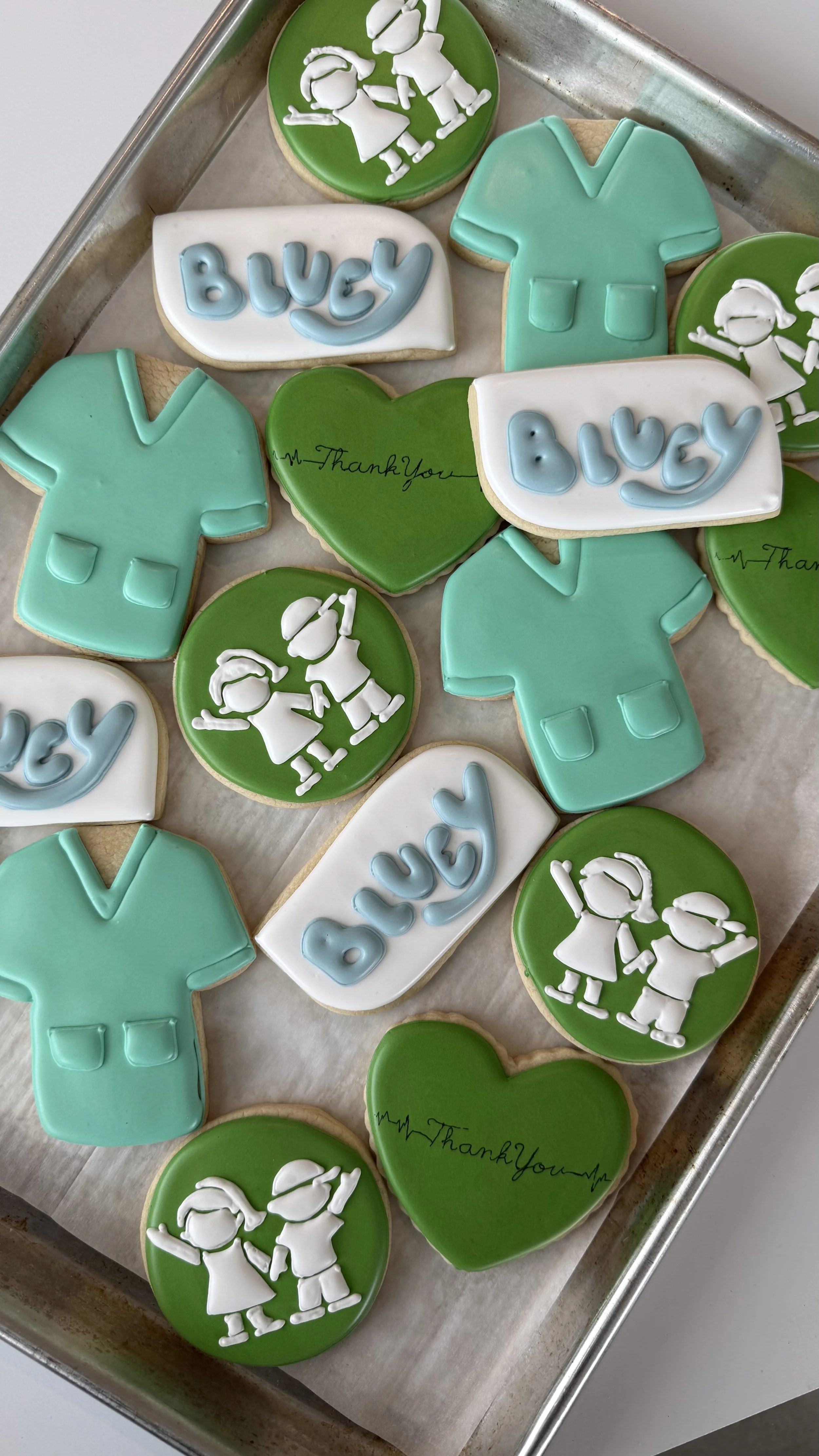 Decorative cookies shaped like scrubs, medical personnel, and hearts with 'Thank You' written on some of the hearts, arranged on a baking tray.