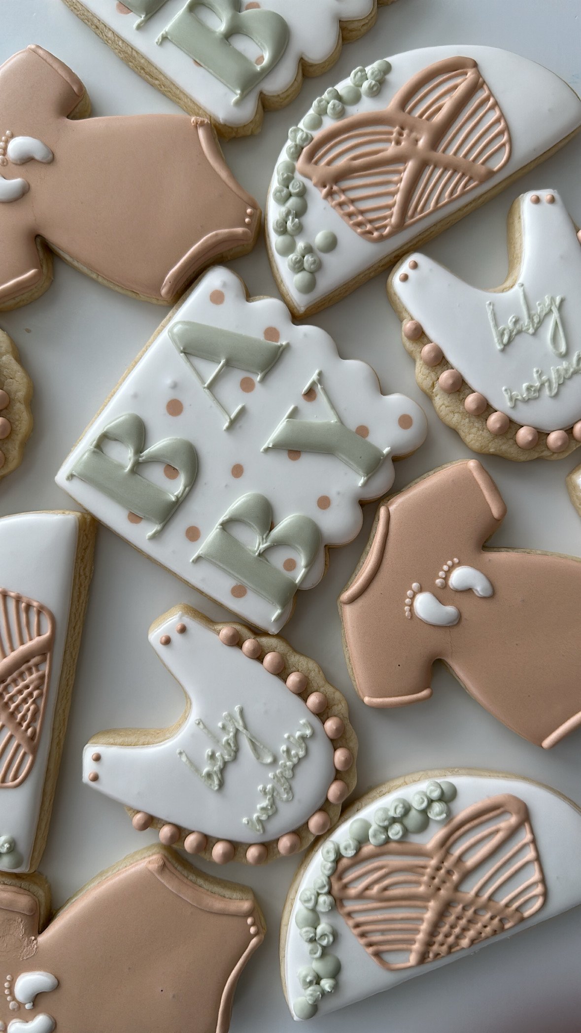 Decorative cookies with neutral pastel pink, white, and green icing, featuring baby-related designs and words like "baby" and "hello baby".