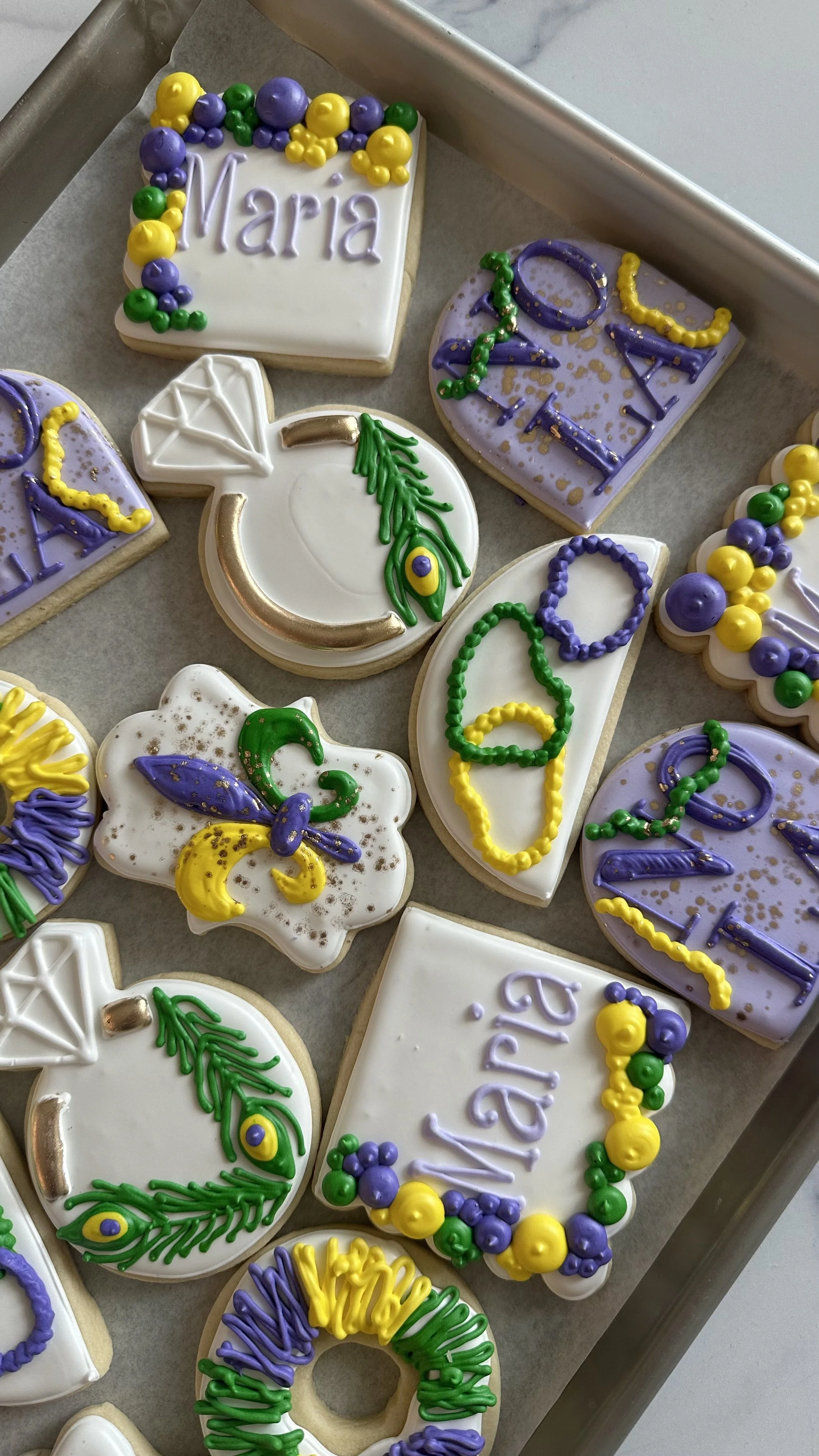 Decorated sugar cookies for Mardi Gras with purple, green, and yellow icing, including the words "Maria" and "2024" and Mardi Gras symbols like beads, peacock feathers, and a diamond.