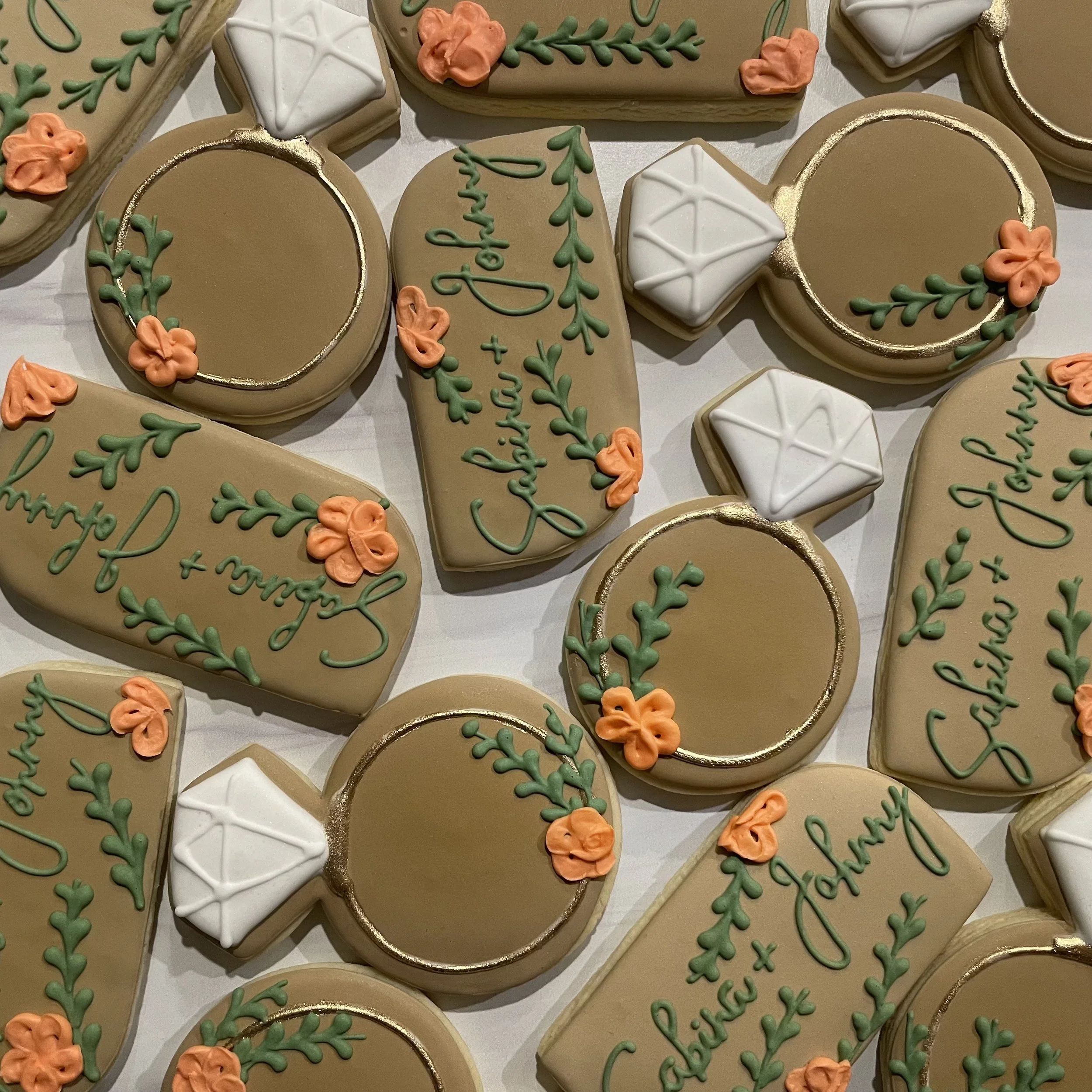 Assorted decorated cookies with white, green, orange icing and gold accents, some with floral and festive designs, arranged on a surface.