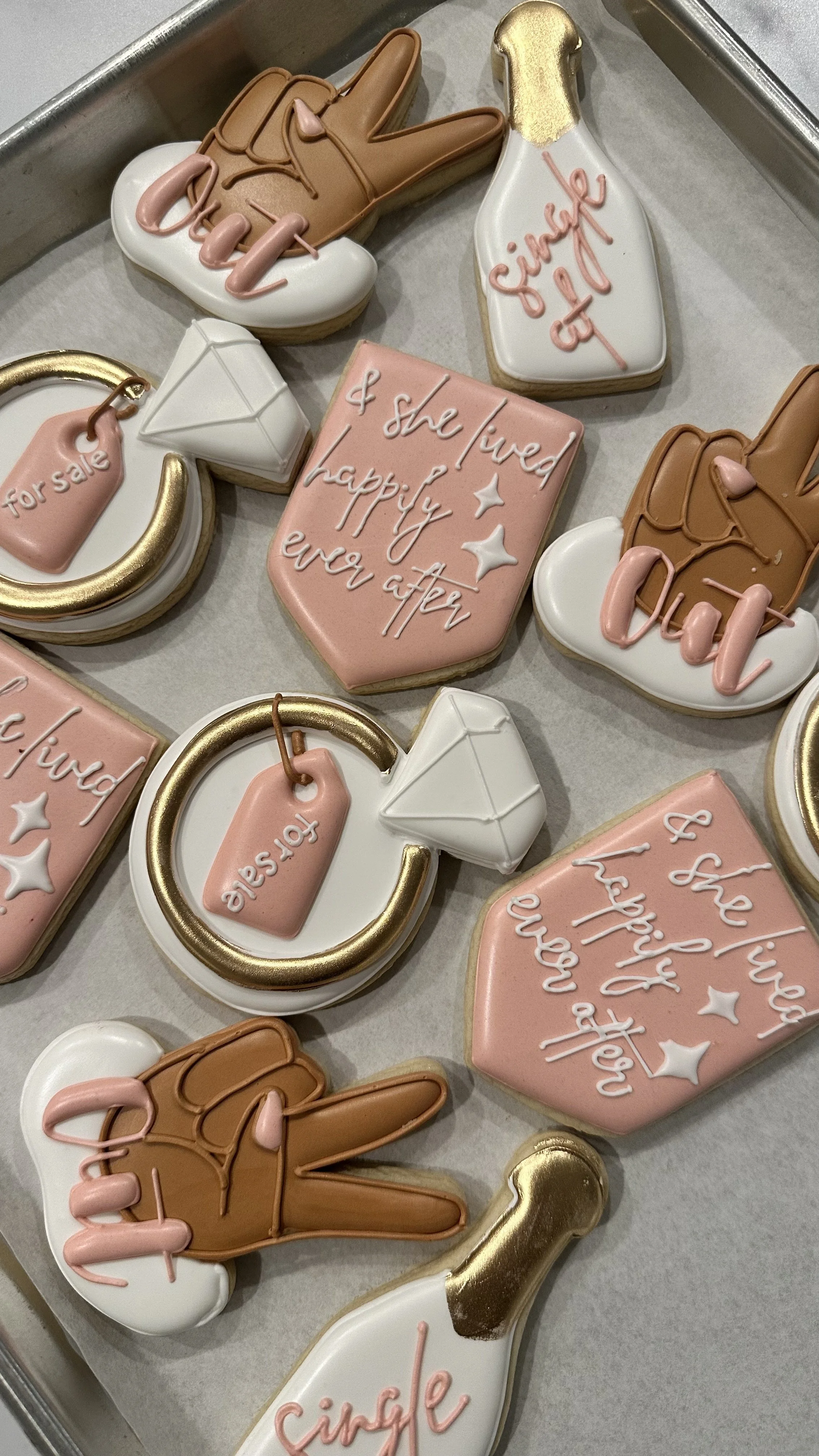 Decorated cookies with messages and symbols related to engagement or marriage, including a diamond ring, a bottle labeled "single" with a ring around it, a pink heart with the message "& she lived happily ever after," and hand-shaped cookies with the