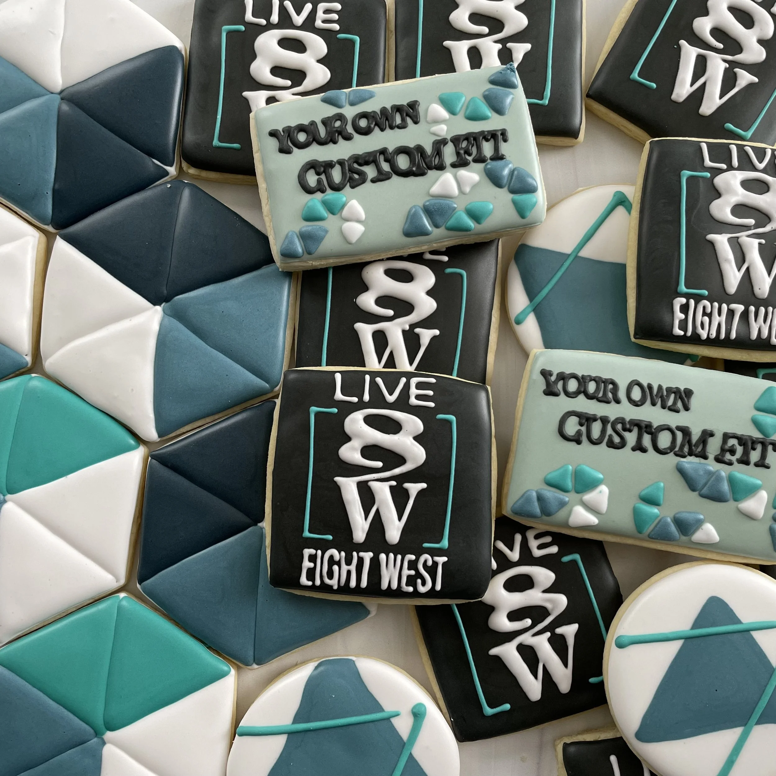 Decorated cookies with inscriptions like "LIVE 8BIT" and "YOUR OWN CUSTOMIZED" in various colors, primarily black, white, and teal, decorated with geometric patterns and sprinkles.