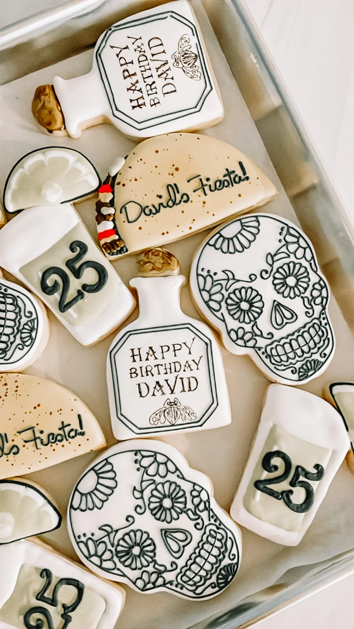 Decorative sugar cookies with black and white Day of the Dead skull designs, some with birthday messages for David, and others with the number 25. Some cookies have inscriptions like "Happy Birthday David" and "David's Fiesta!".